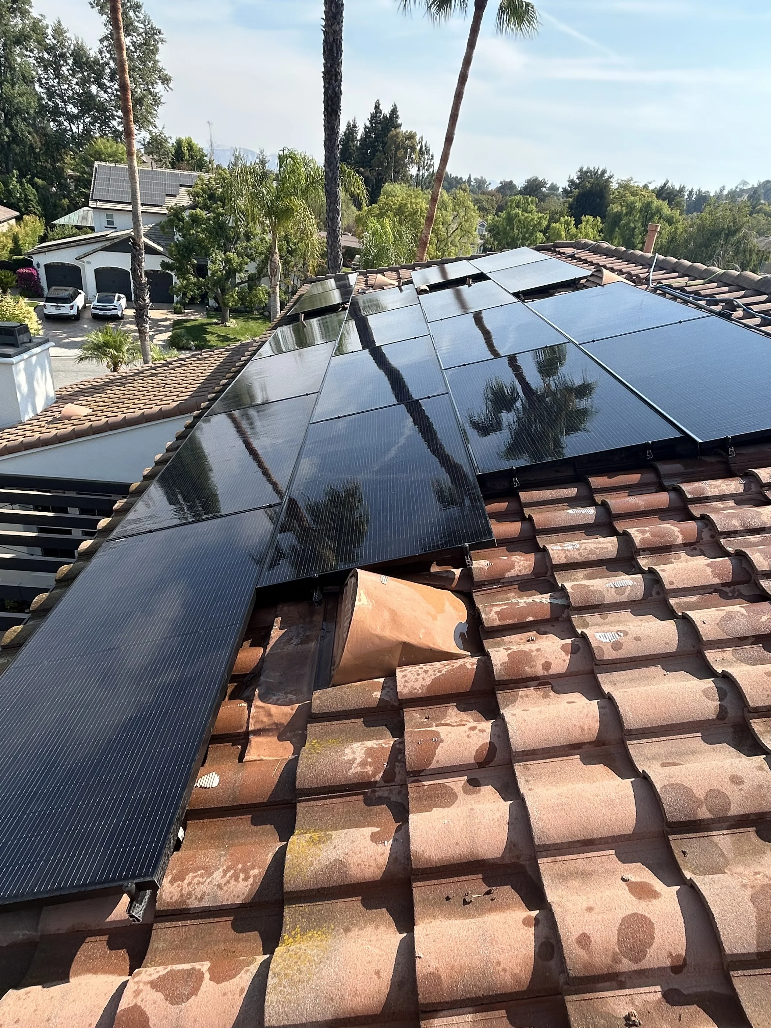 Solar panels installed on a tiled roof of a house with trees, other houses, cars, and a driveway in the background.