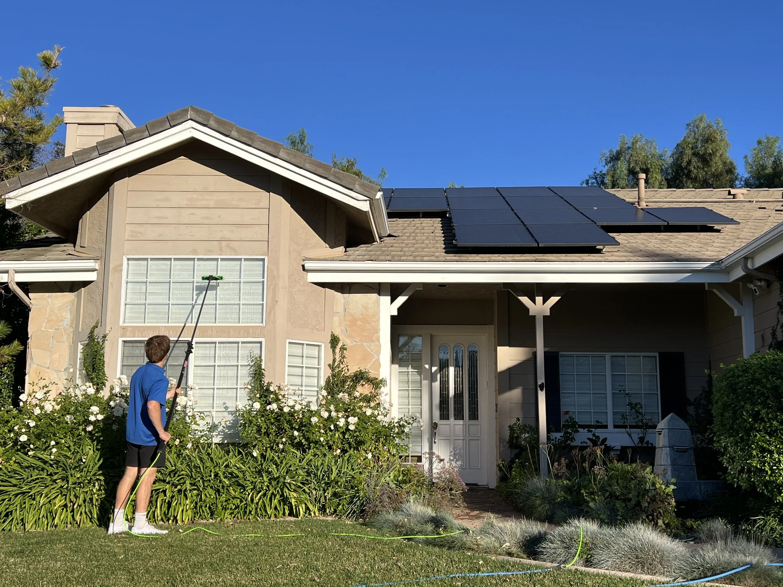 A person using a pole to clean solar panels installed on the roof of a house. The house has beige siding, large windows, and a garden with plants and flowers in the foreground. The sky is clear and blue with trees in the background.