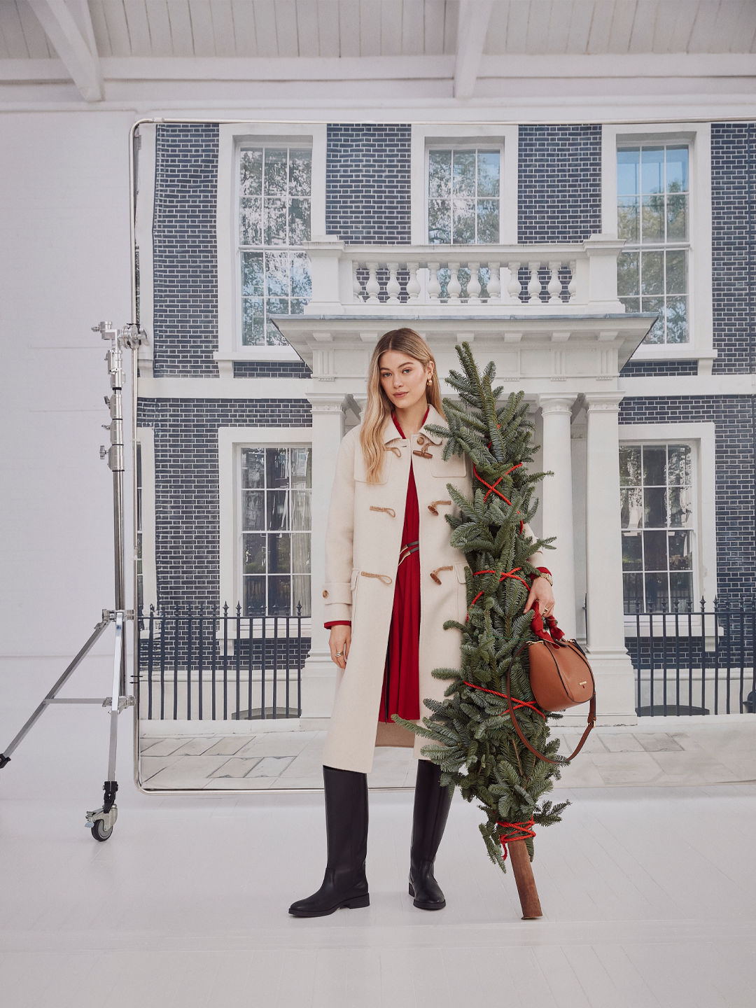 Mulher com cabelo loiro e vestido vermelho, usando casaco bege e botas pretas, segurando uma árvore de Natal decorada com fitas vermelhas em frente a uma fachada de casa grande com janelas brancas e detalhes em branco.