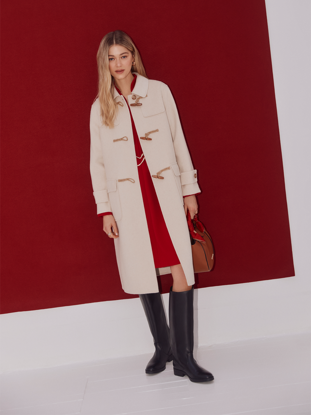 Mulher vestida com casaco bege, vestido vermelho e botas pretas, segurando uma bolsa marrom, posando contra fundo vermelho.