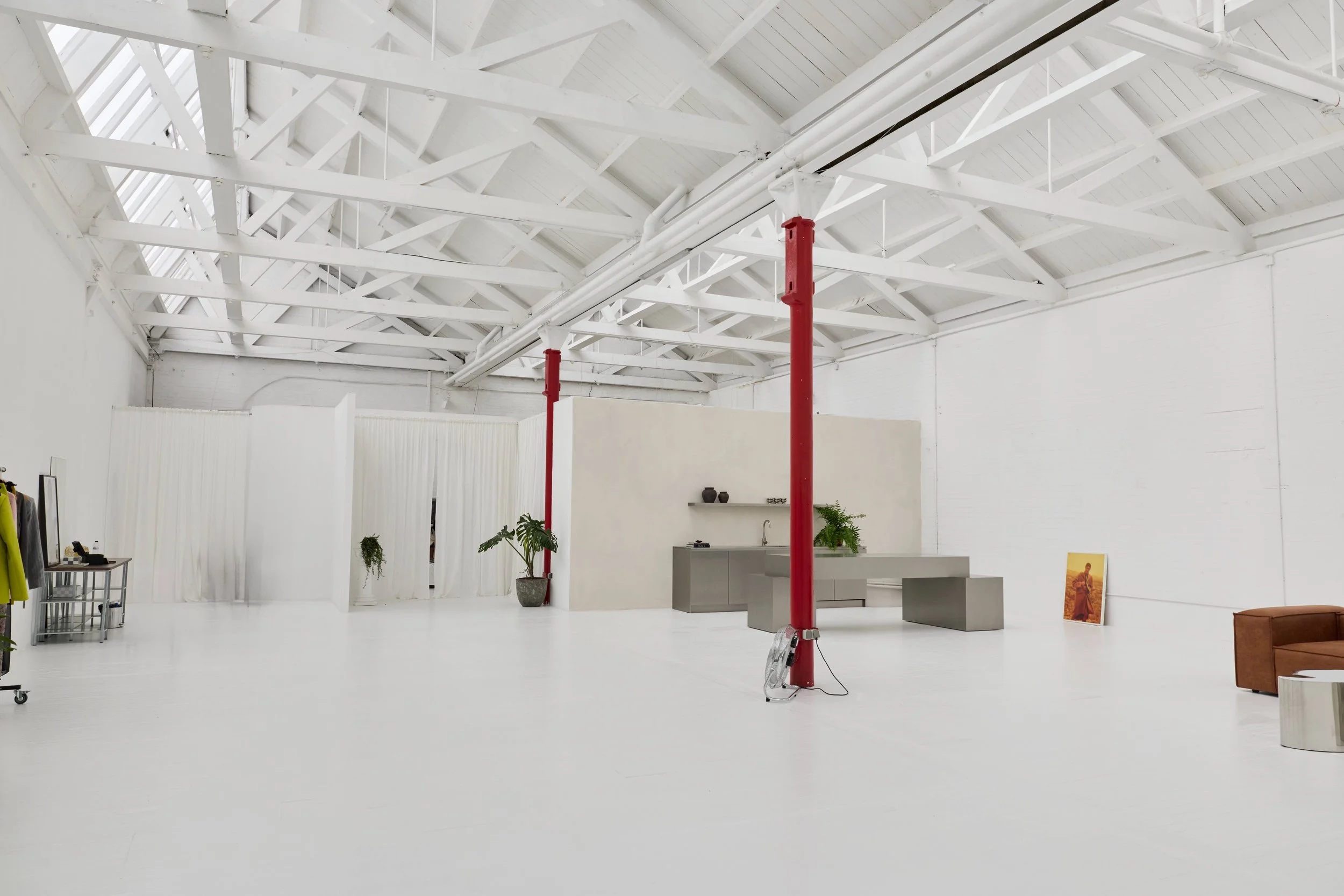 Espaço amplo e minimalista com teto de estrutura de vigas brancas e paredes brancas. Há uma cozinha com balcão cinza, algumas plantas, uma pintura apoiada no chão, uma cadeira de couro marrom, um espelho e uma mesa de apoio com roupas e objetos.