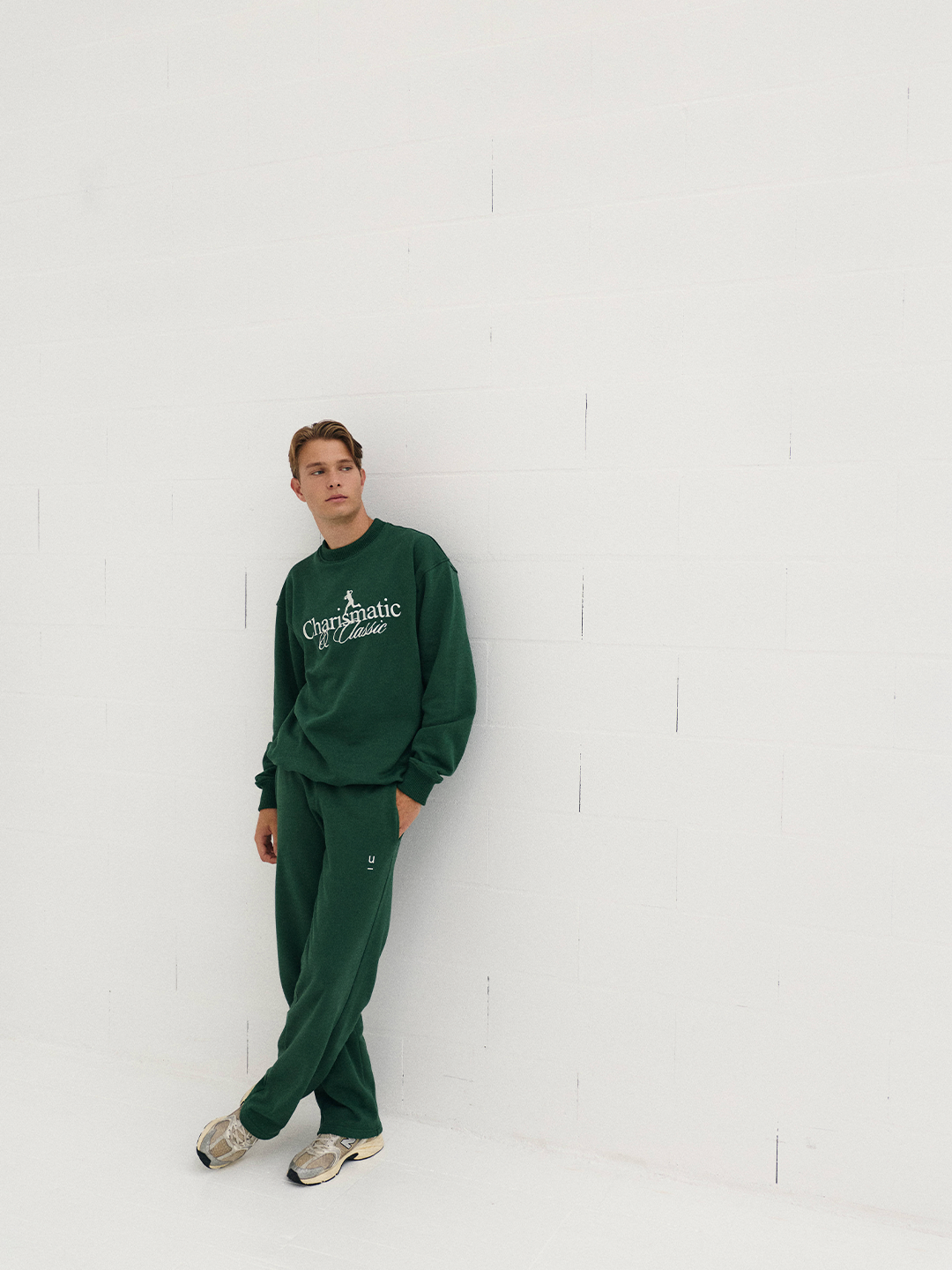 Jovem vestindo roupa esportiva verde, de pé contra uma parede branca, olhando para o lado.