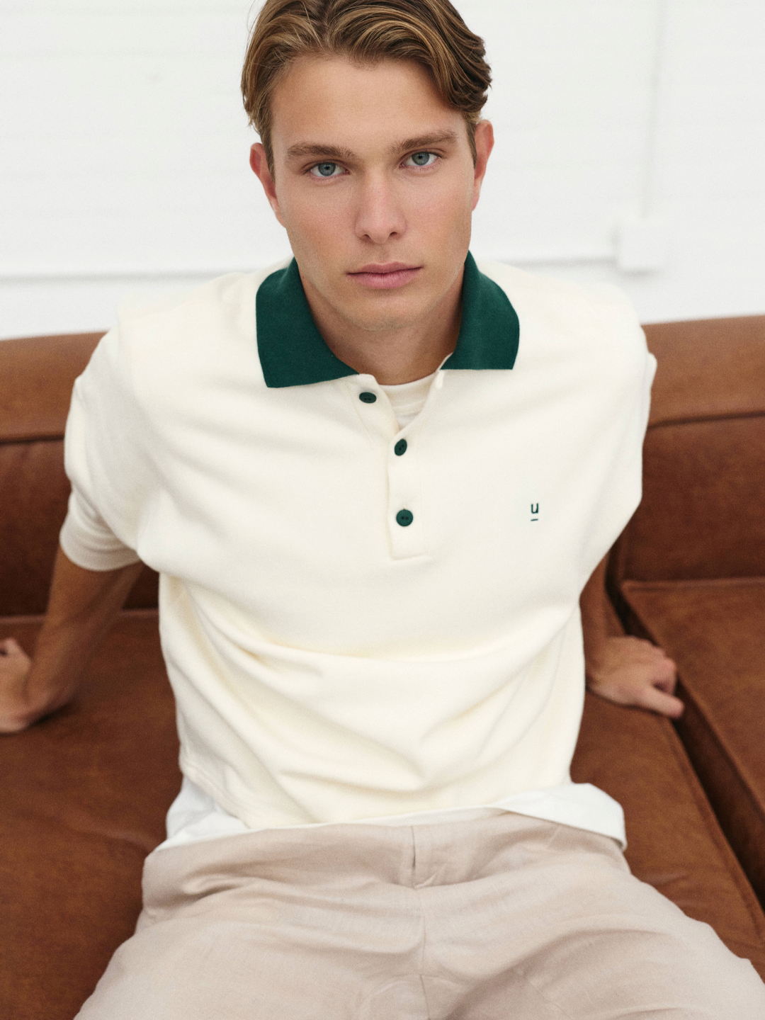 Homem de cabelo castanho claro e olhos azuis, usando camisa polo branca com gola verde, sentado em um sofá de couro marrom.