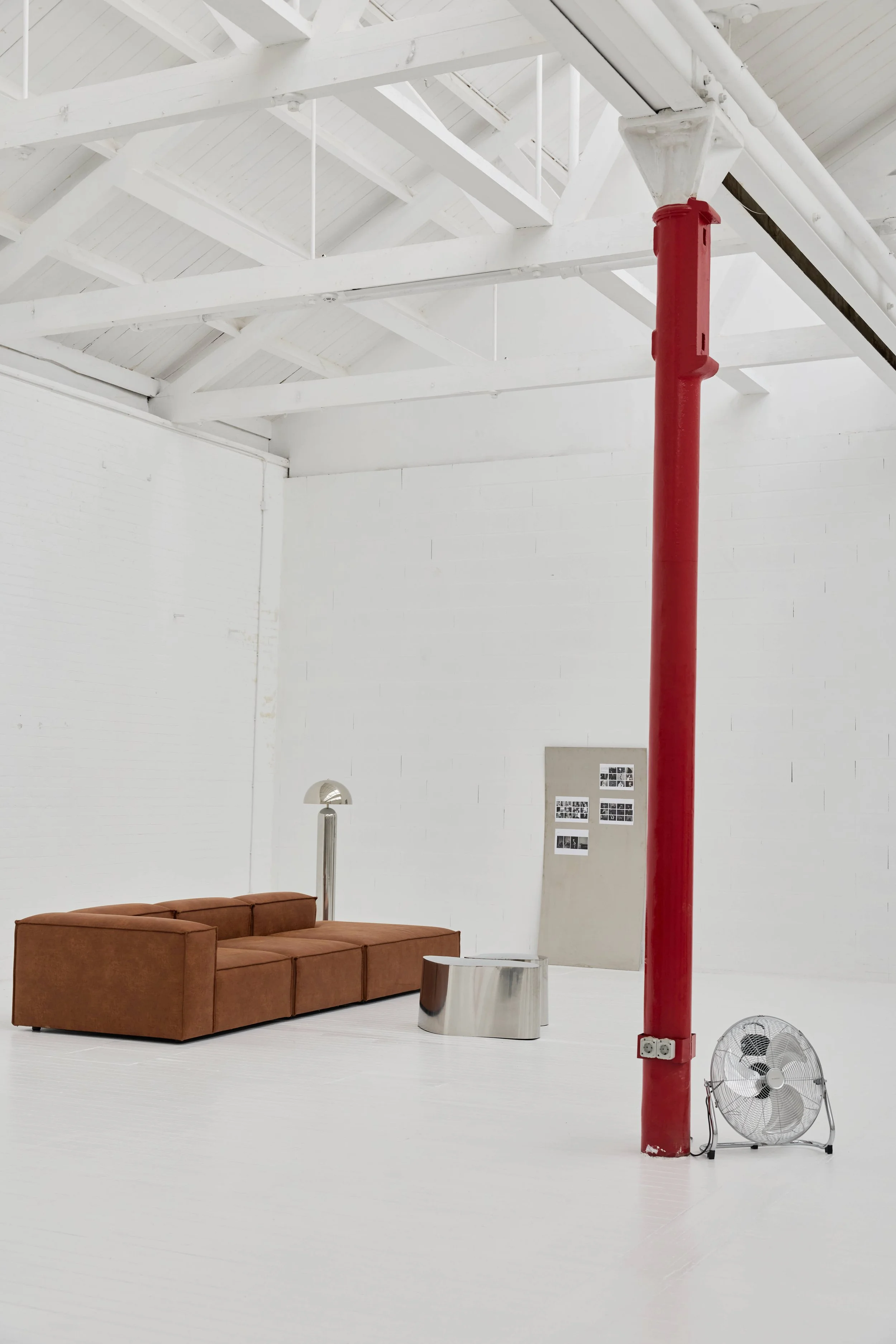 Sala com sofá marrom, lâmpada de pé, painel de fotos, mesa de metal, ventilador e uma coluna vermelha.