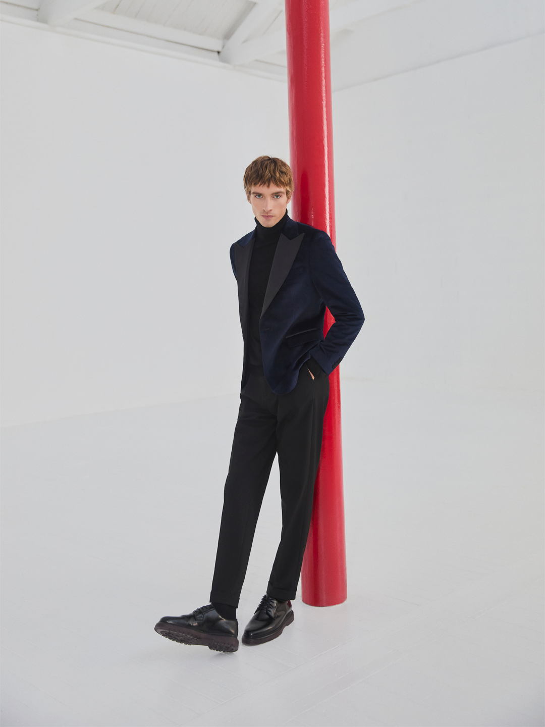 Homem jovem com cabelo castanho claro, vestindo terno preto, camisa preta de gola alta, blazer azul escuro, calçando sapatos sociais pretos, apoiado em uma coluna vermelha em um espaço branco minimalista.