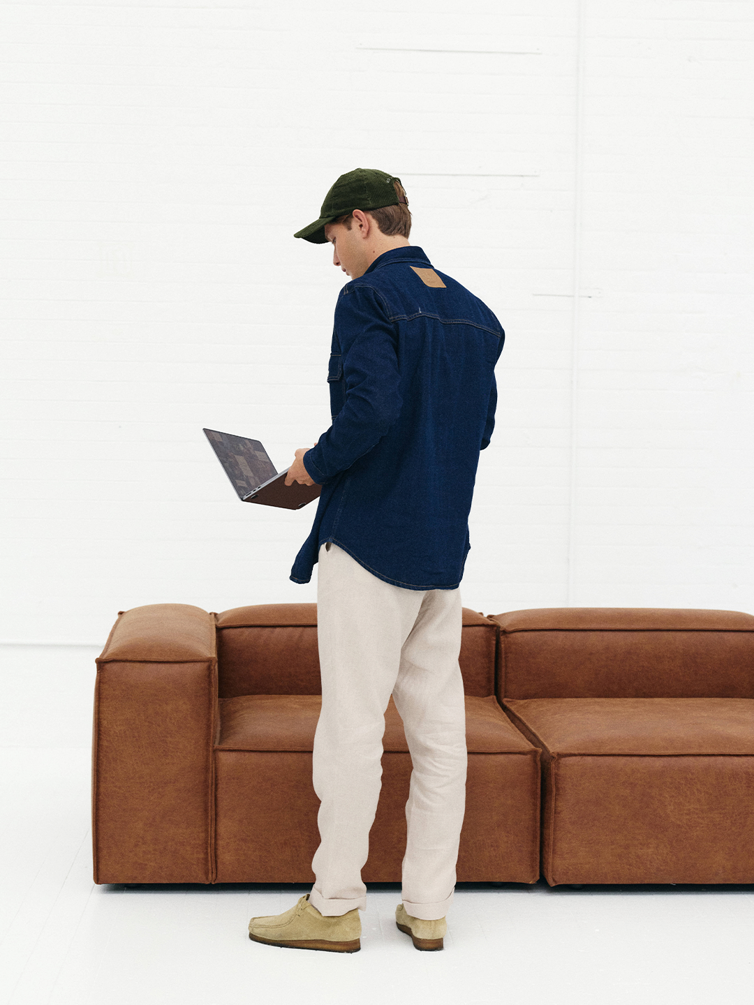 Homem de peitoral médio, usando boné verde, jaqueta jeans azul, calça clara e sapatos bege, segindo um laptop em um ambiente minimalista com sofa marrom e paredes brancas.