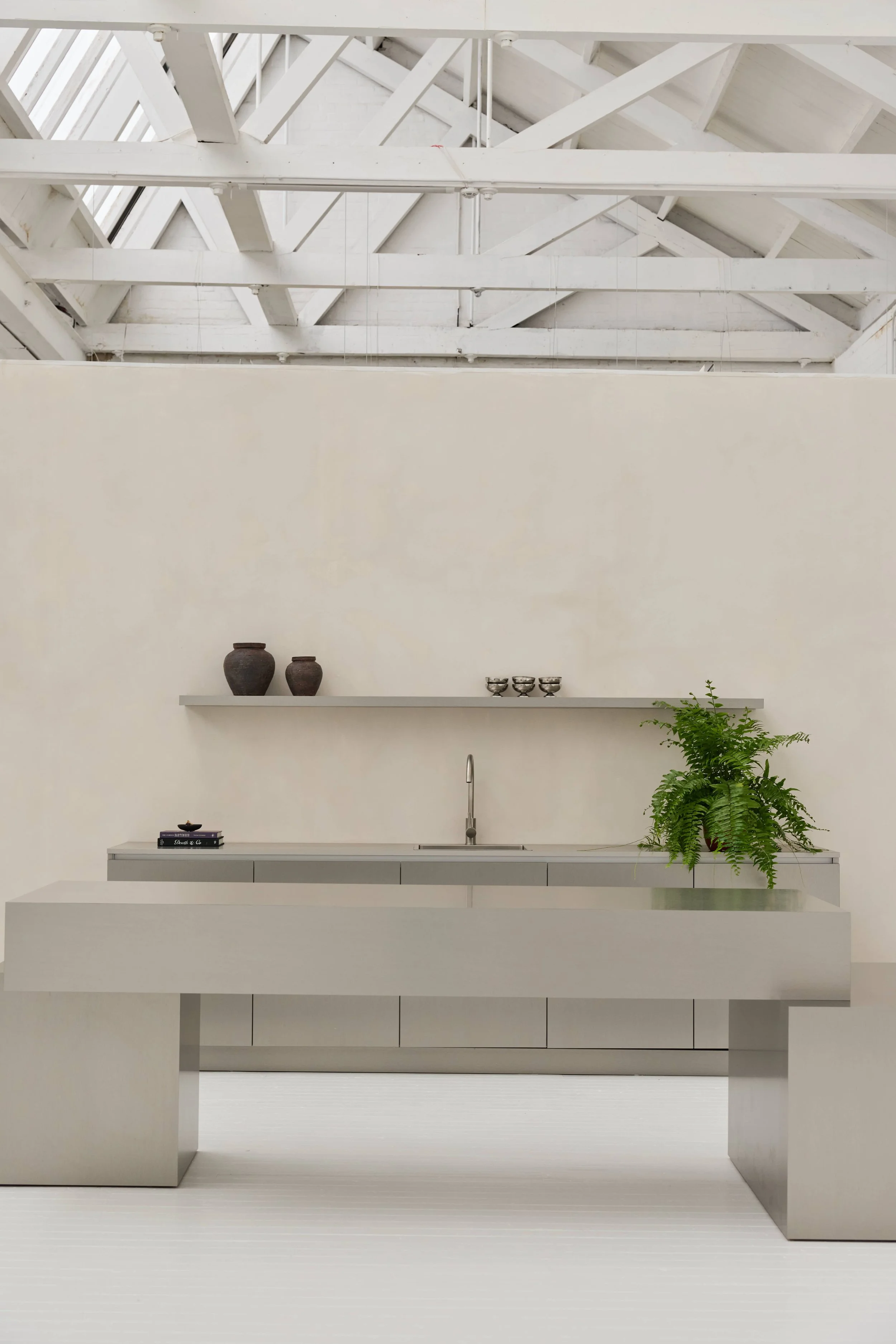 Cozinha minimalista com bancada e prateleira cinza, vaso de plantas e objetos decorativos, parede branca e teto de estrutura de madeira branca.