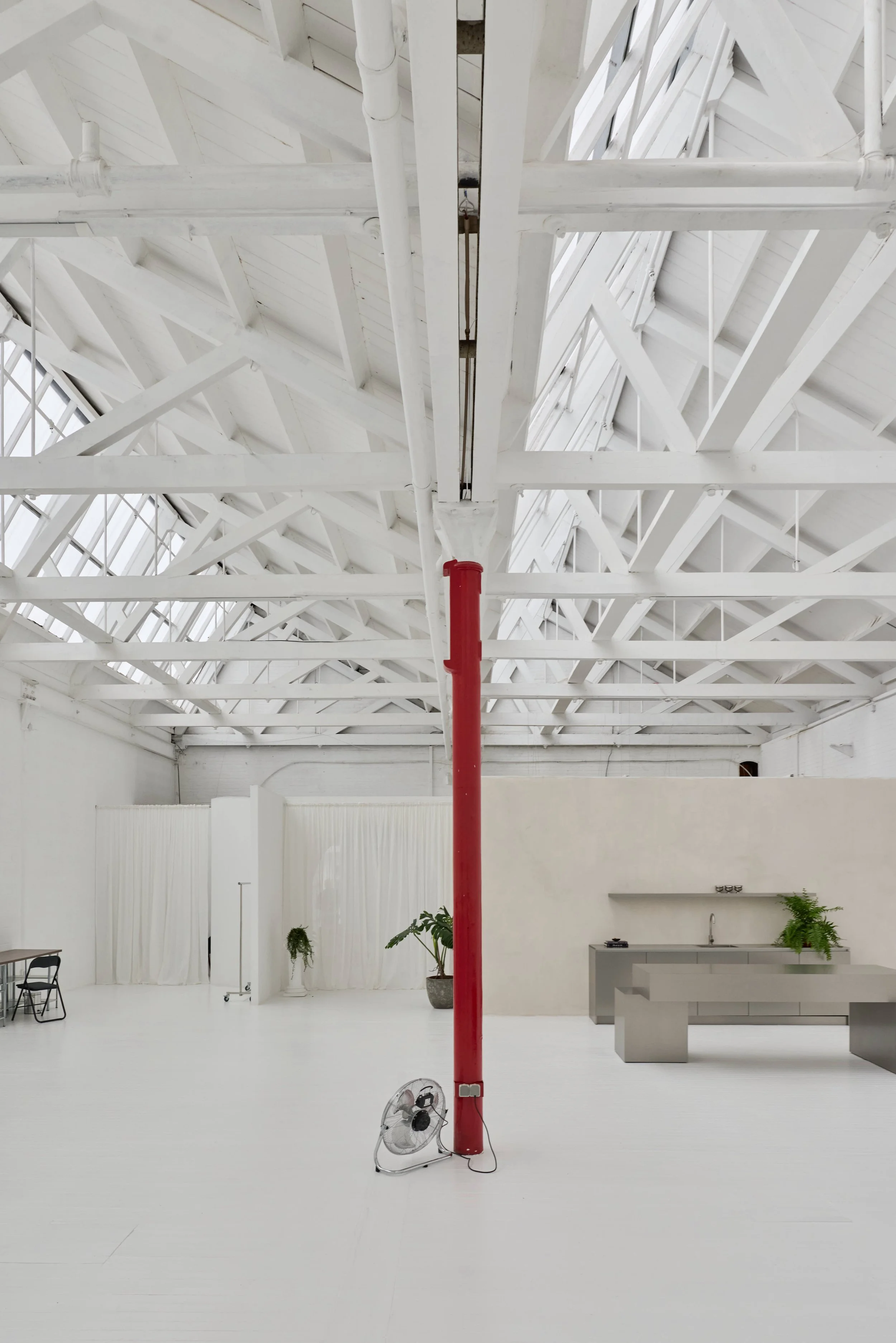 Interior de um espaço vazio com teto de estrutura de madeira pintada de branco, uma coluna vermelha central, plantas verdes, bancadas de concreto e uma fan coil no chão. Toda a decoração é predominantemente branca.