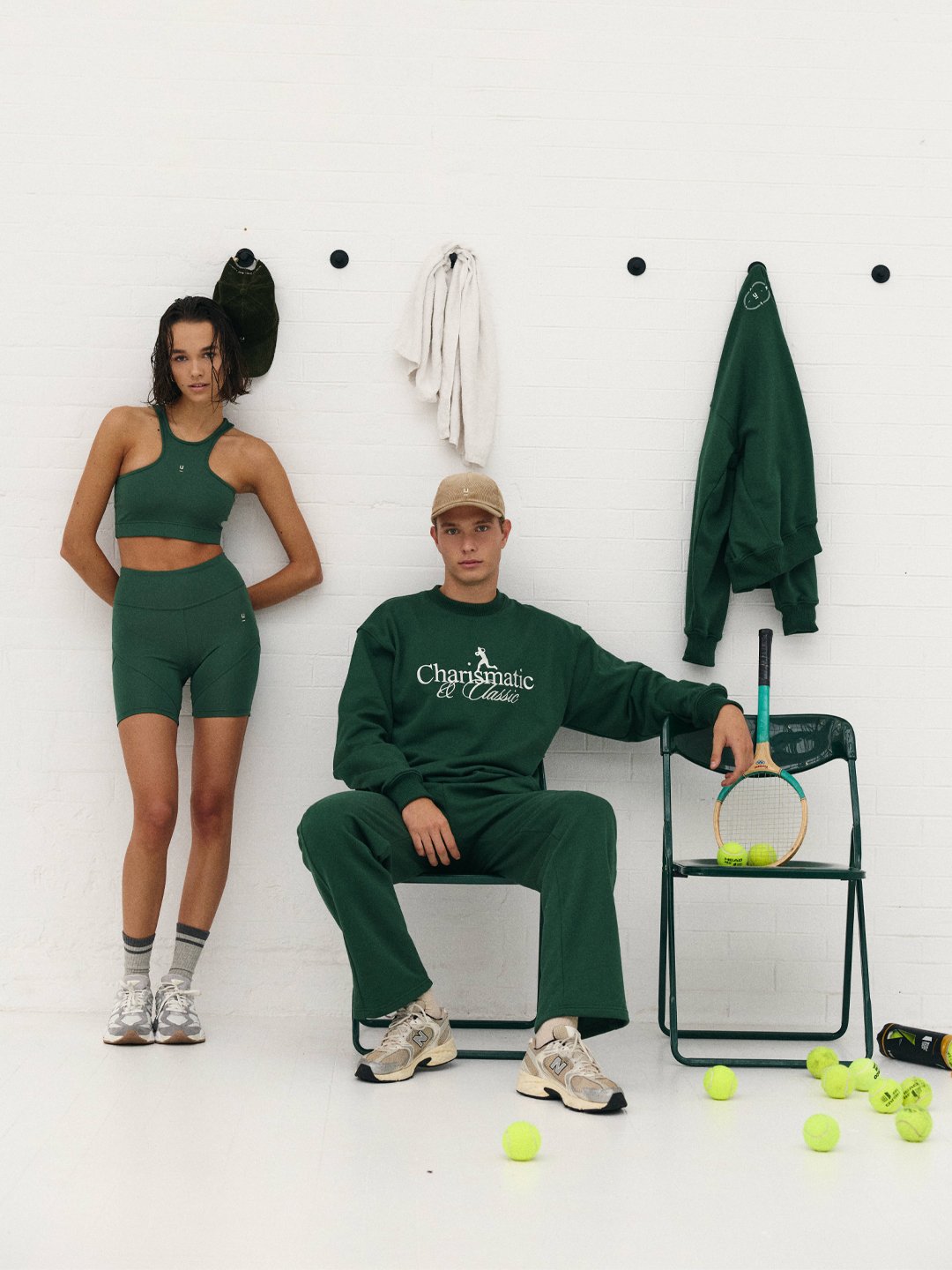 Homem sentado em cadeira com roupa esportiva verde, mulher de roupa similar em pé ao lado, com raquete e bolas de tênis ao redor numa parede branca