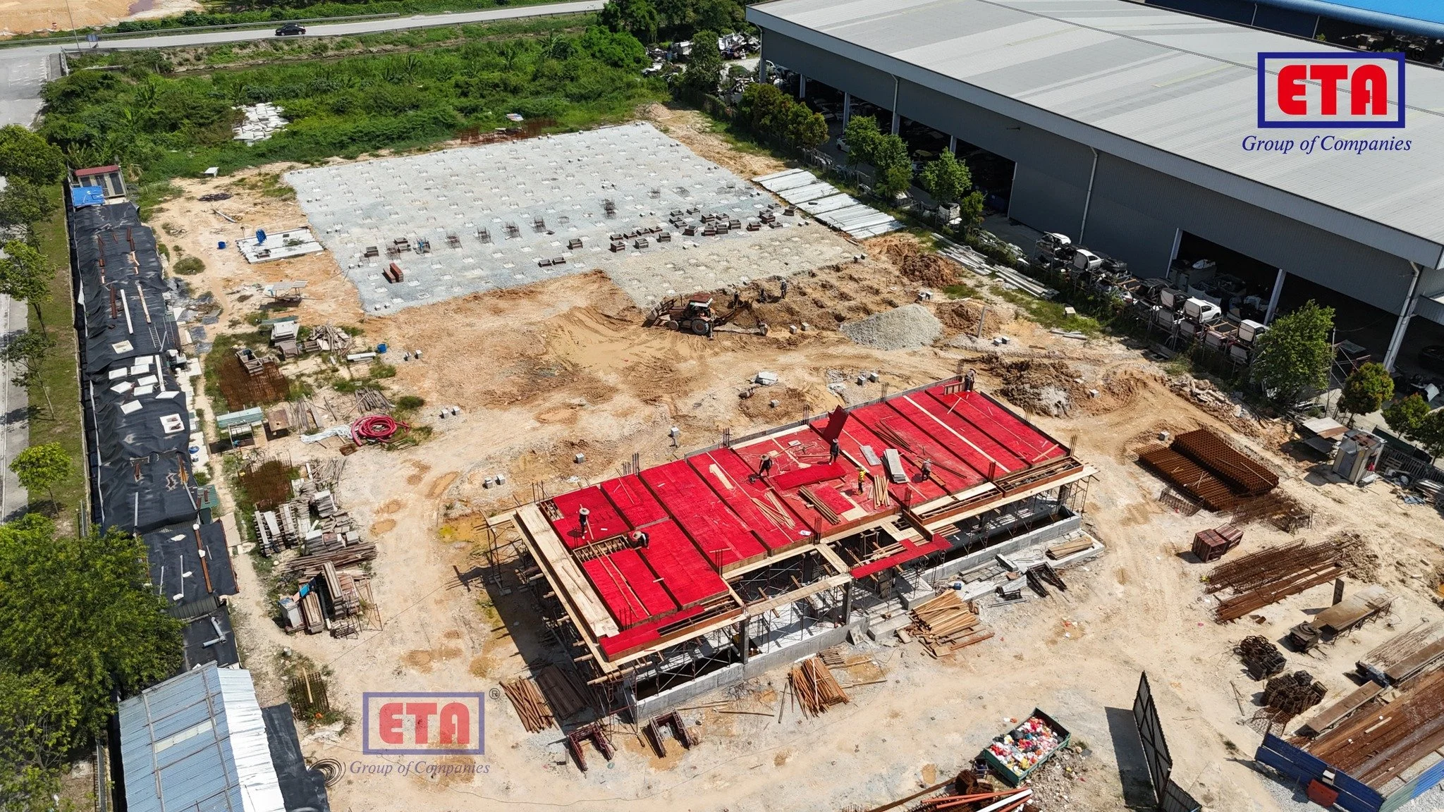Aerial view of a construction site showing a partially built building with red flooring, construction workers, scattered building materials, and machinery surrounded by greenery and nearby structures.