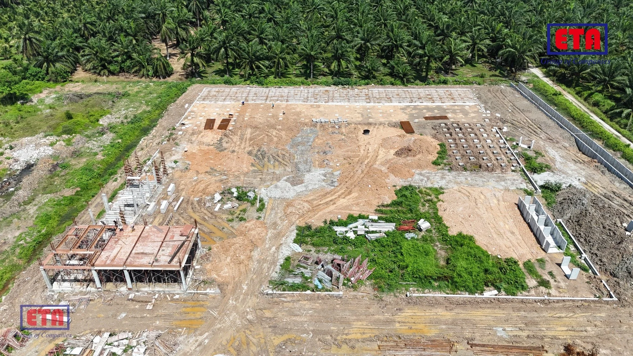 Aerial view of a construction site with partially built structures, scattered construction materials, and greenery in the background, featuring palm trees and a fenced perimeter.