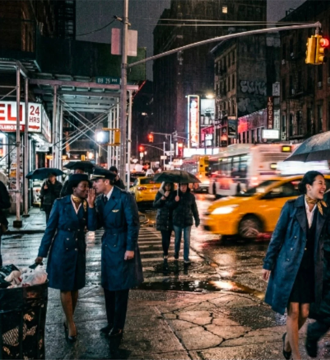 Nighttime city street scene with people holding umbrellas, yellow taxis, and blurred moving vehicles with illuminated signs in the background.