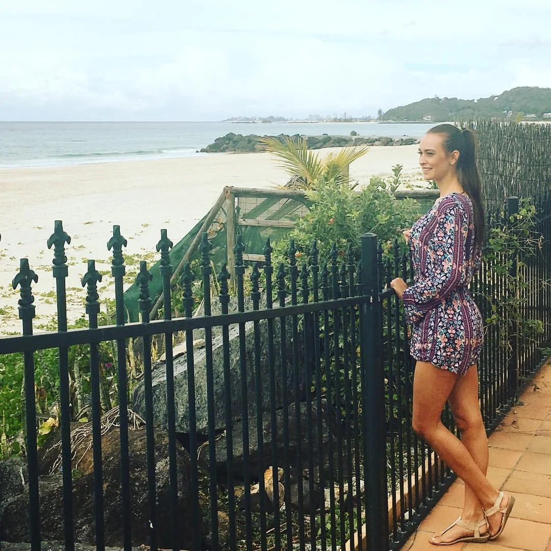 A woman with long dark hair smiling and standing by a black metal fence at the beach, wearing a patterned romper and sandals, with the ocean, sandy beach, and distant hills in the background.