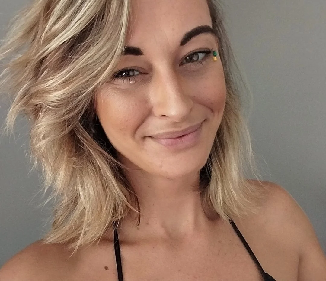 Close-up of a smiling woman with wavy blonde hair, light makeup, and a small tattoo on her collarbone, wearing a black spaghetti strap top.