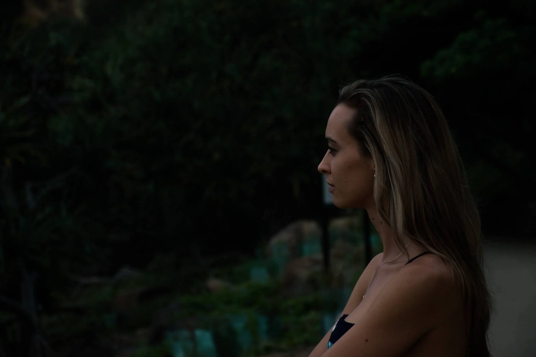 A woman with long hair standing outdoors near a body of water and green foliage.