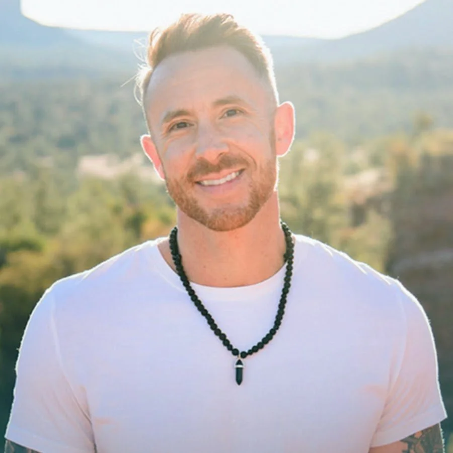 Smiling man with light skin, short styled hair, and a beard, wearing a white t-shirt and a black beaded necklace with a pendant, standing outdoors with mountains and trees in the background.