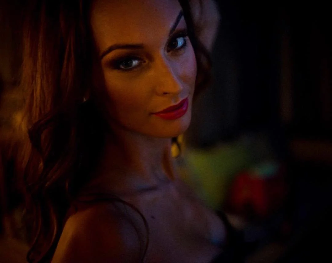 A woman with long auburn hair and red lipstick looking at the camera in a dimly lit environment.