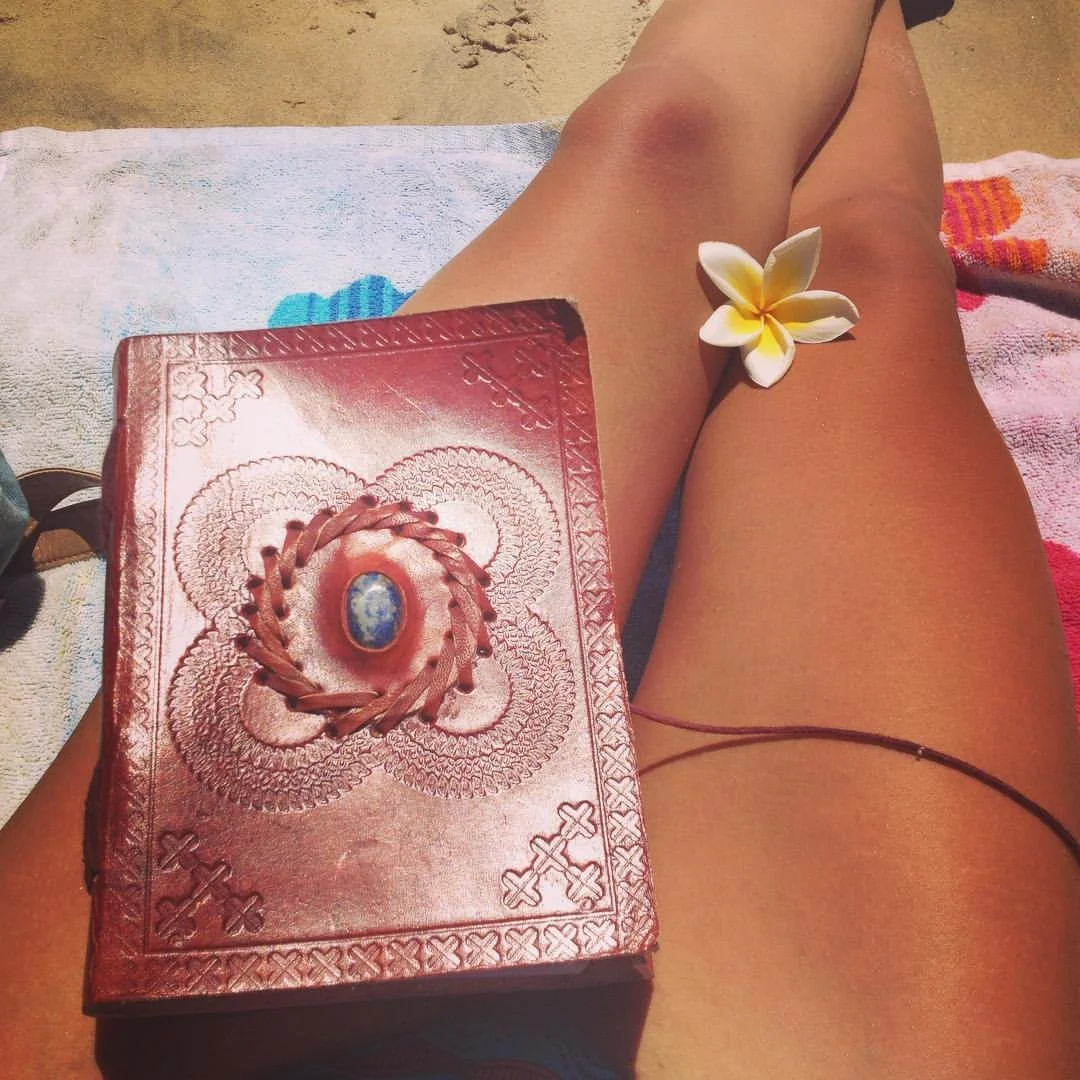 A person sitting on a towel at the beach with a red book, a white and yellow fangapani flower, and part of a black necklace visible.
