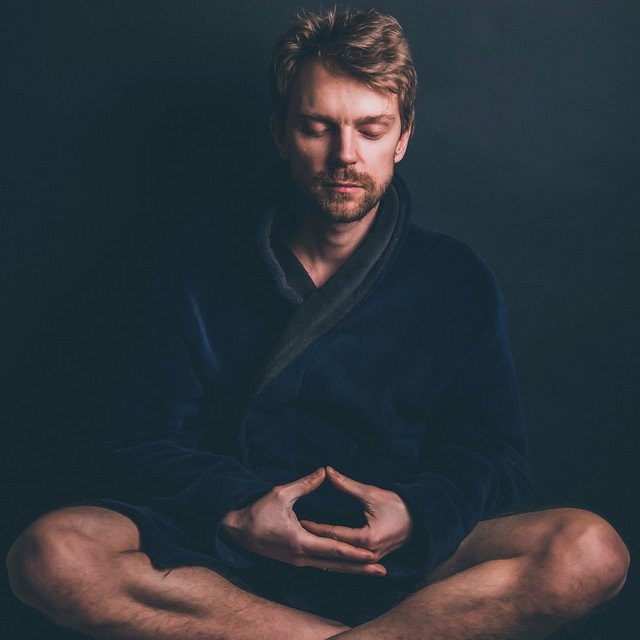 A man sitting cross-legged with closed eyes, practicing meditation, wearing a dark robe against a dark background.