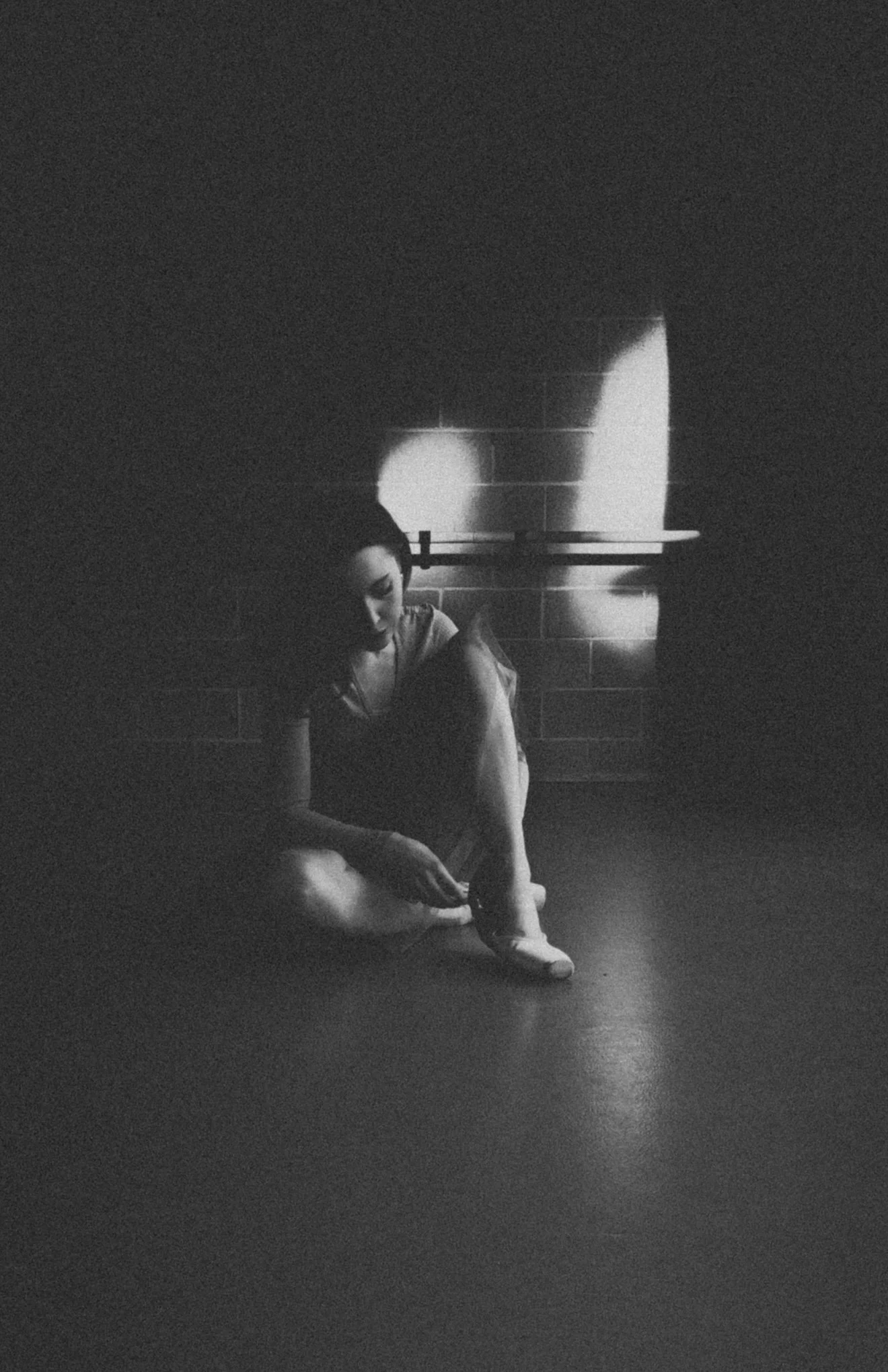 A woman sitting on the floor in a dimly lit room, adjusting her ballet shoe, with a shadow of her face cast on the wall behind her.