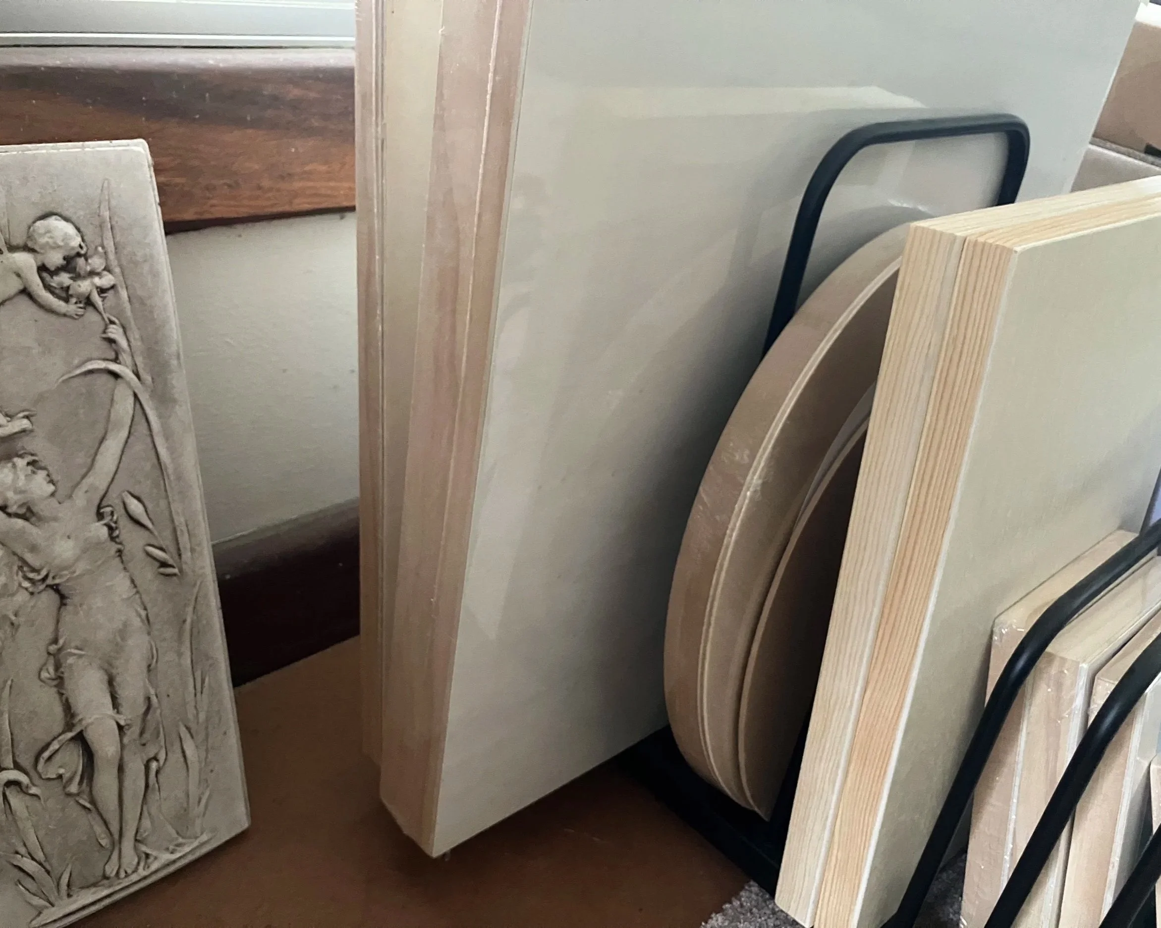 Various artists' canvas and table art frames stored vertically, including a carved relief sculpture, flat canvas boards, and rounded canvas shapes, leaning against a black metal stand.