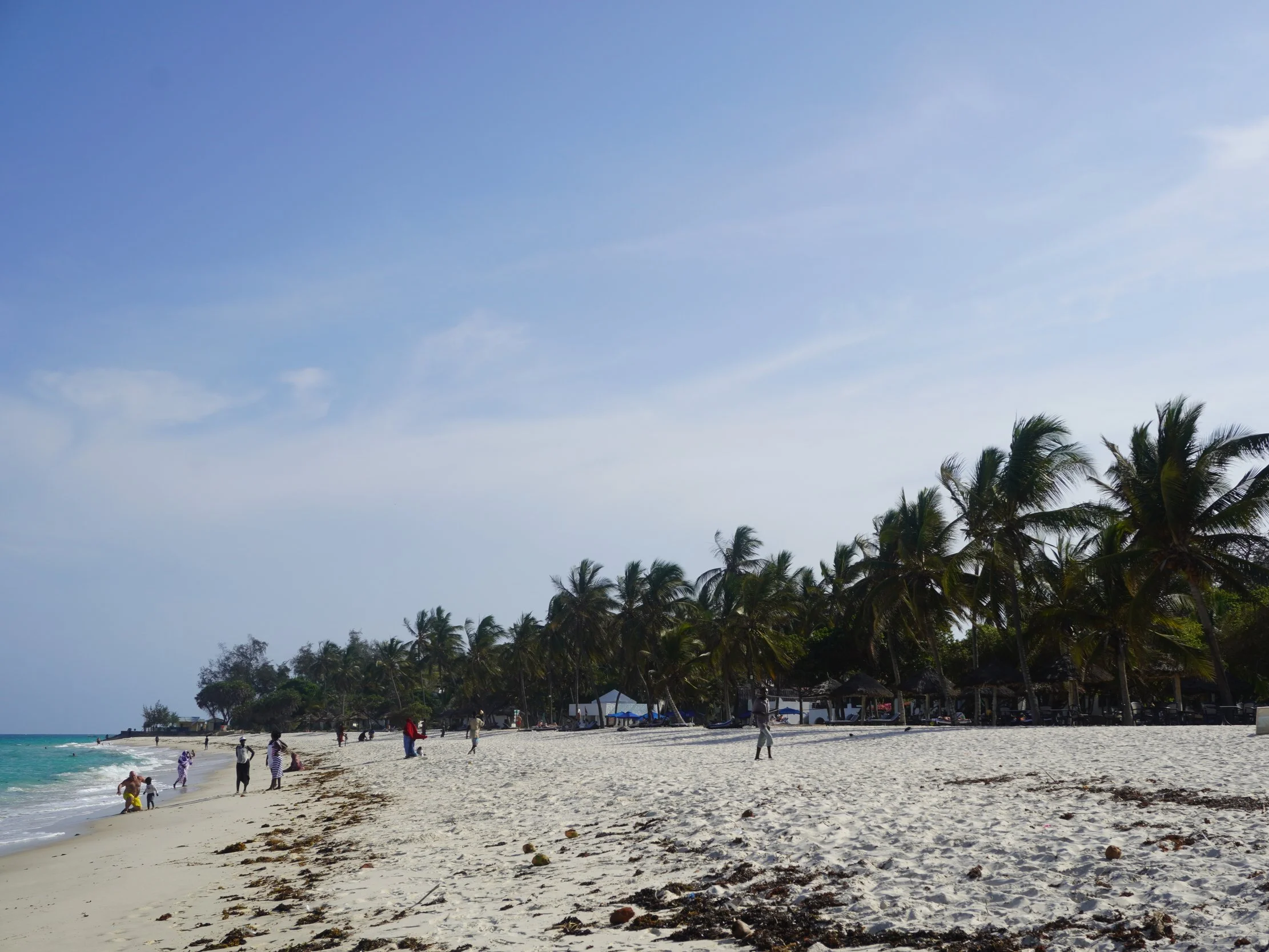 Beach with white sand, palm trees, and several visitors walking near the shoreline under a partly cloudy sky.