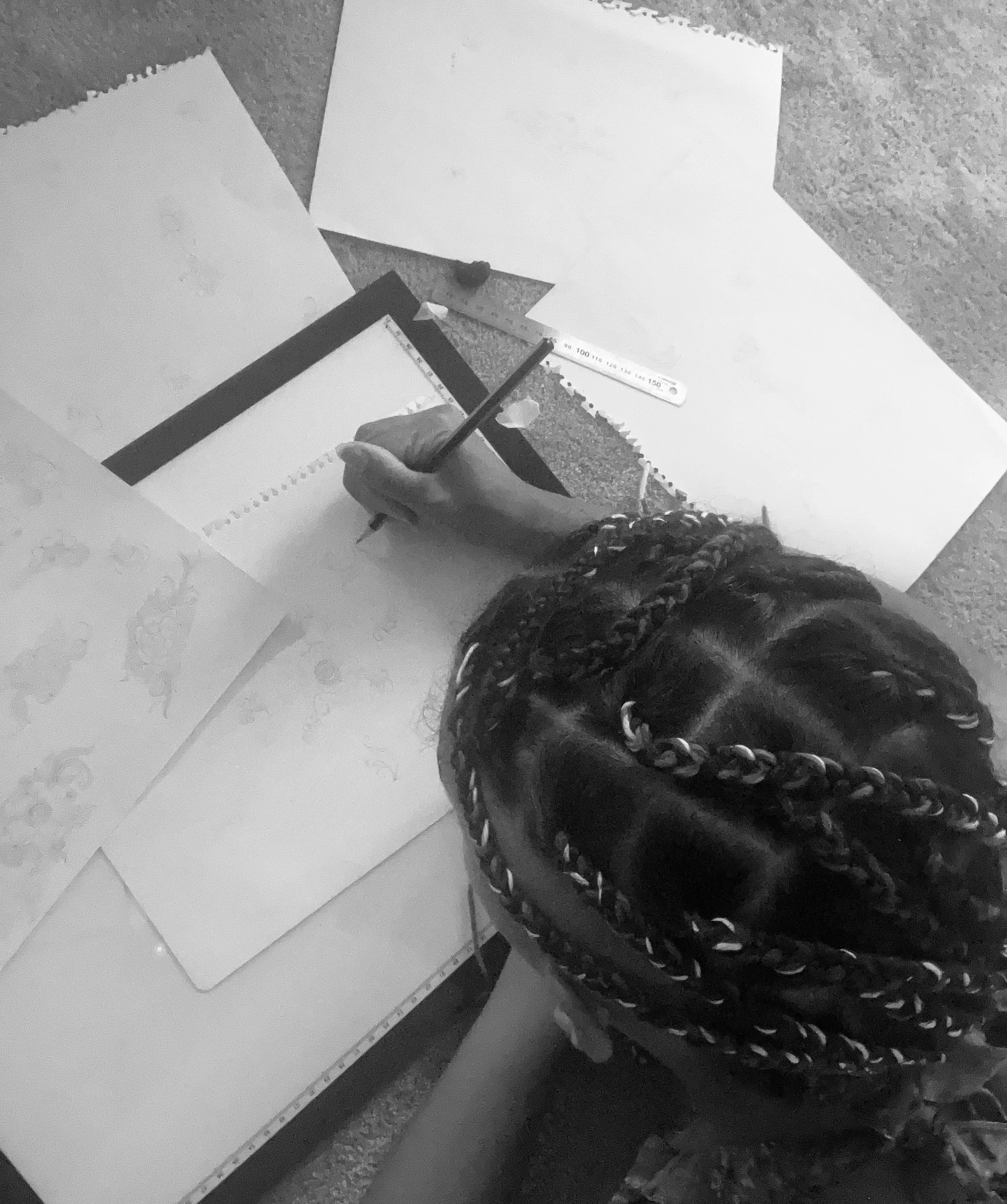 A person with braided hair is drawing on a piece of paper with a pen, surrounded by other sheets of paper, a ruler, and a pencil on a textured surface.