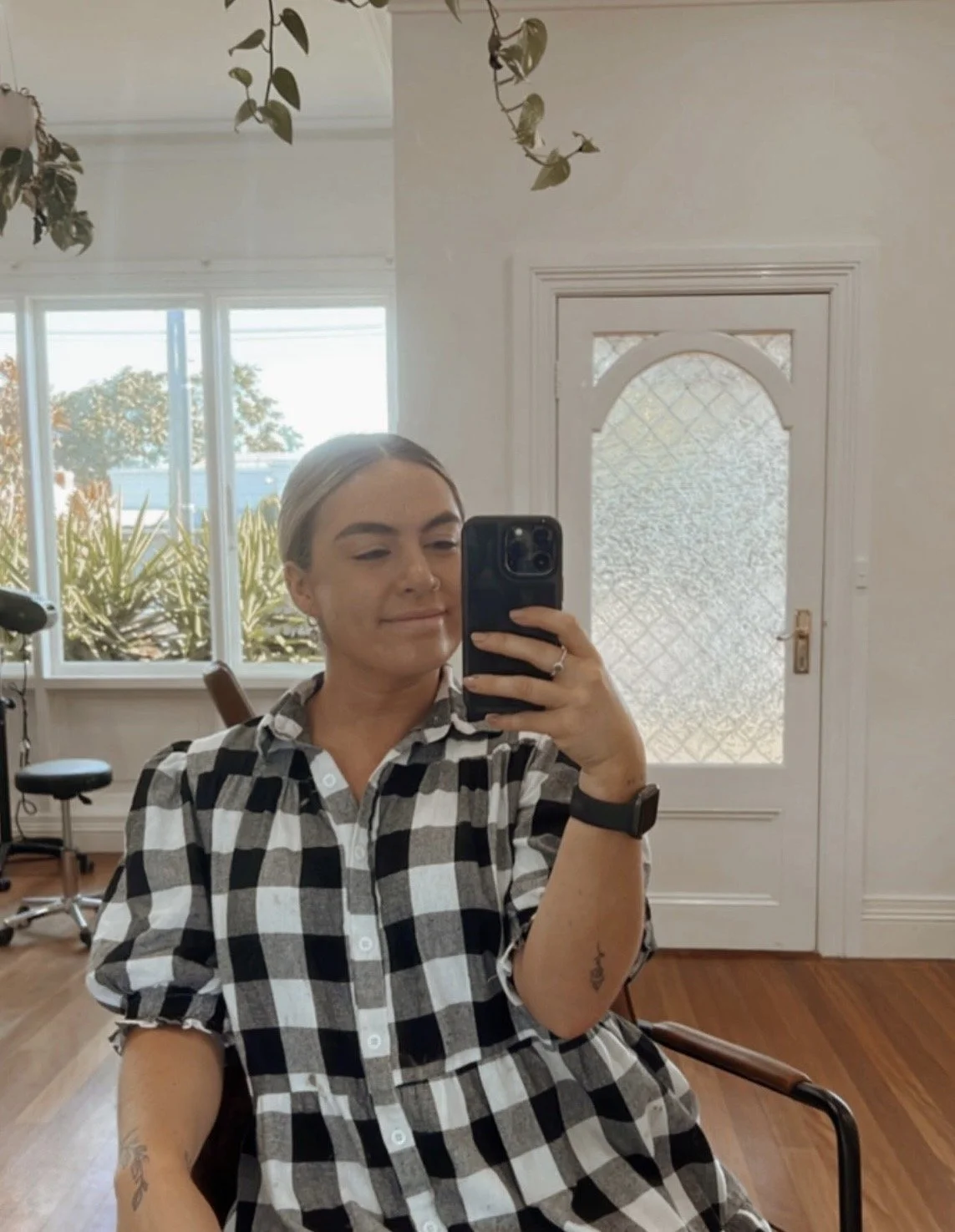 A woman taking a selfie with a smartphone in a room with a large window and a door with frosted glass. She is smiling, wearing a black and white checkered dress, a smartwatch, and has tattoos on her arms.