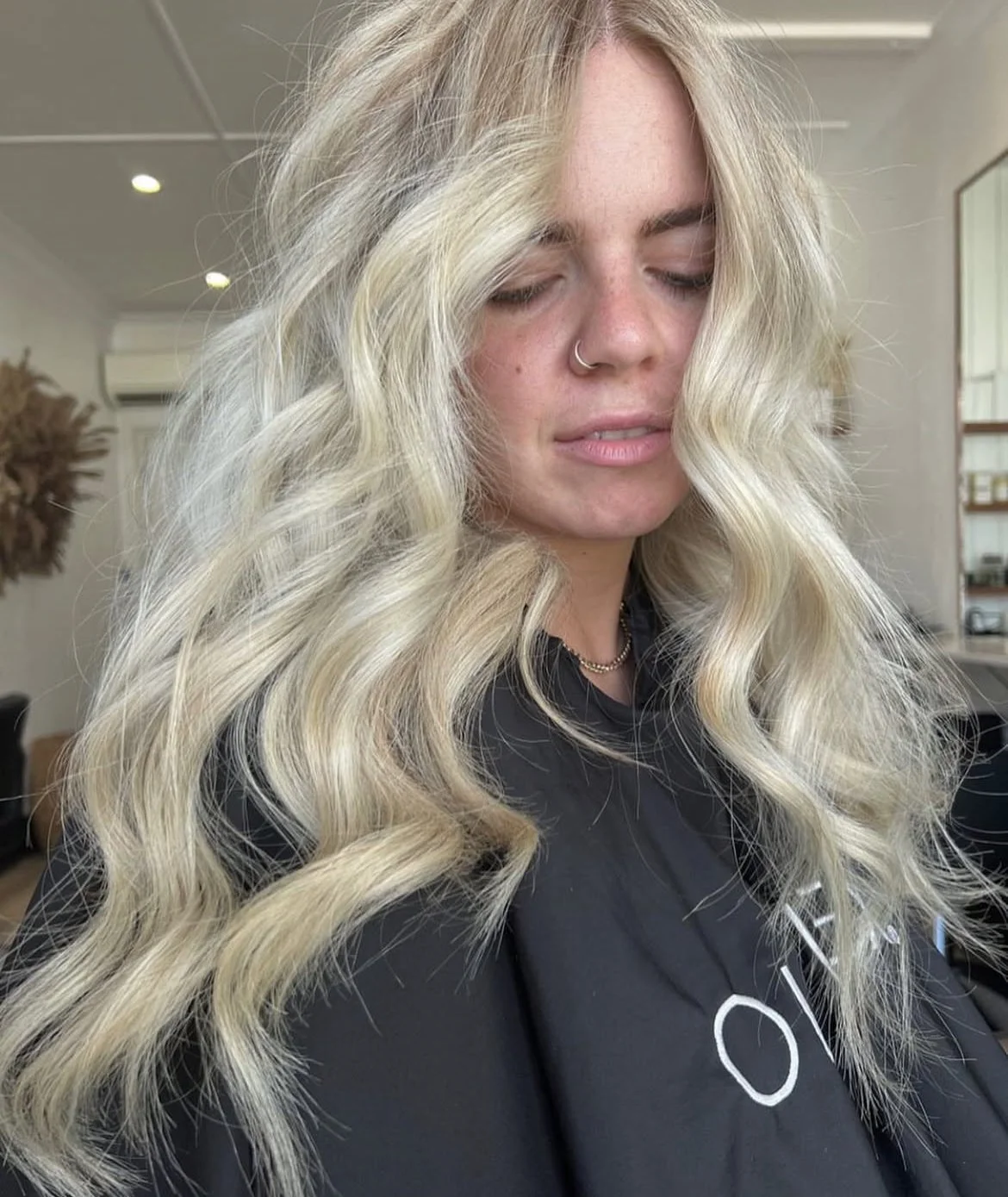 A woman with long, voluminous blonde hair with loose waves, wearing a nose ring, a black shirt, and layered necklaces, sitting indoors with her eyes closed.