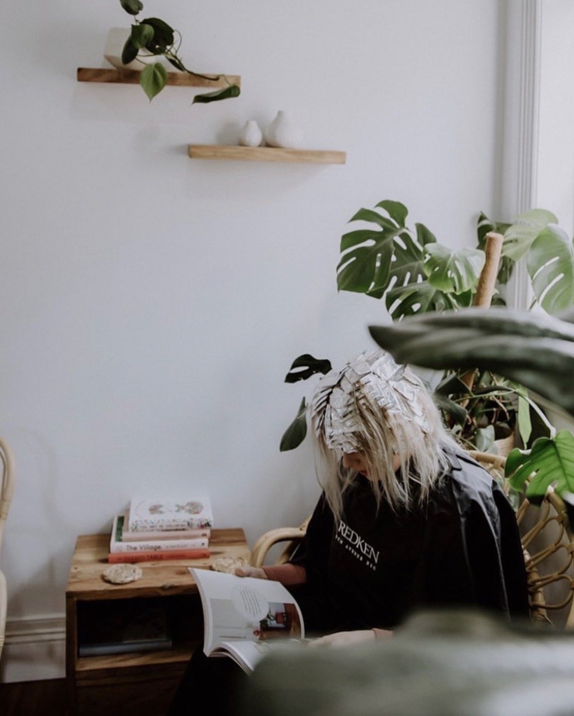 A person with platinum blonde hair reading a magazine while sitting in a room with green plants and wooden furniture.