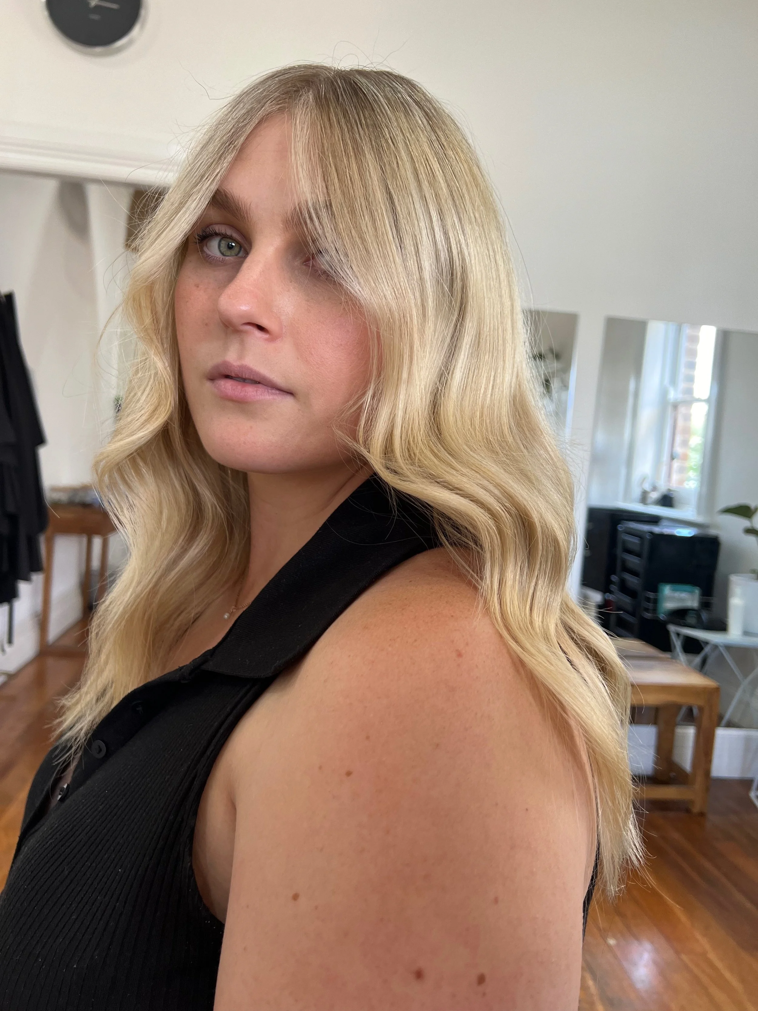 A woman with blonde wavy hair and fair skin is looking at the camera, wearing a black sleeveless top, in a bright room with large windows and wooden furniture.