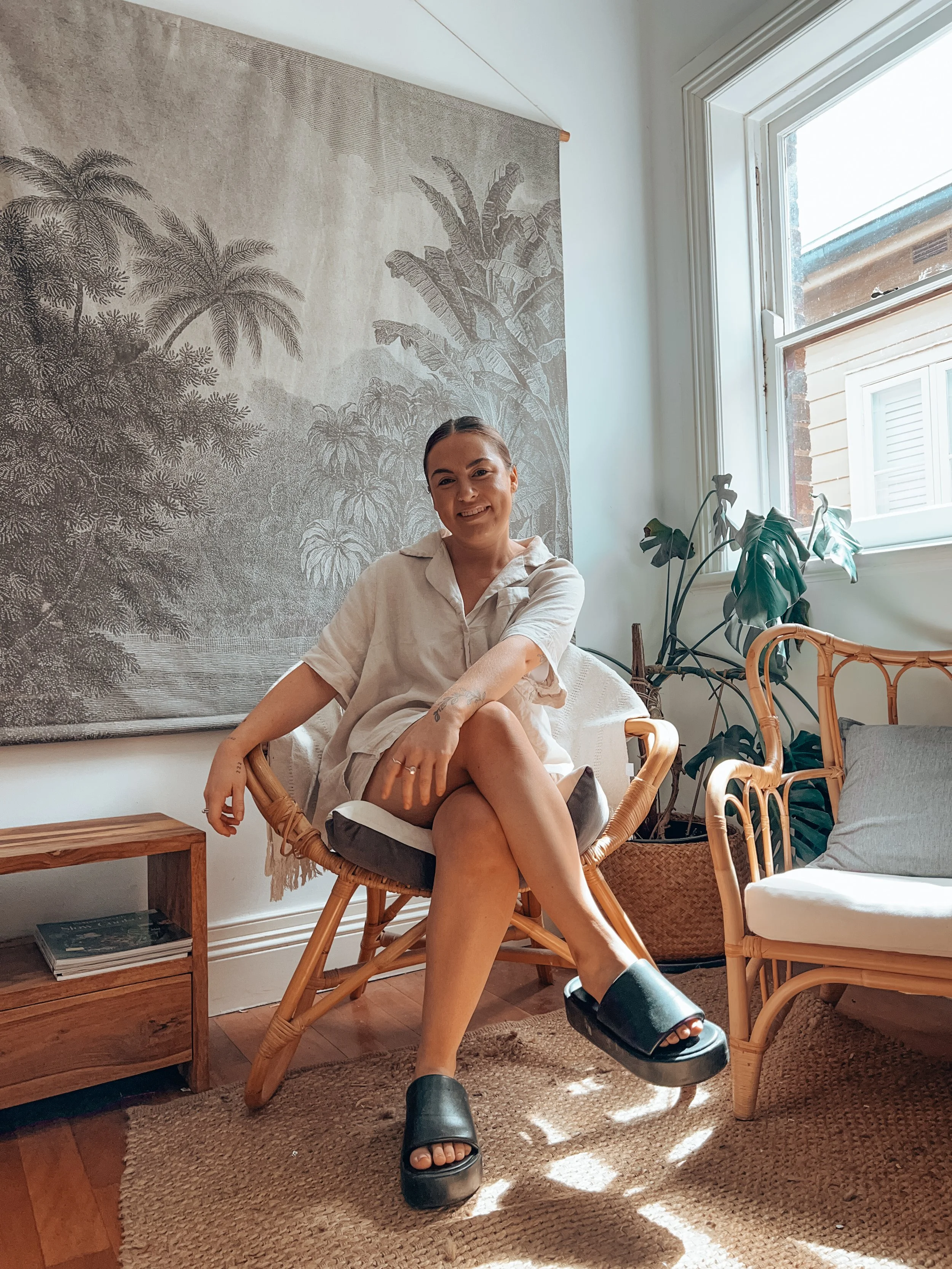 A woman sitting on a rattan chair in a cozy, sunlit room with greenery and a tapestry of jungle scene in the background.