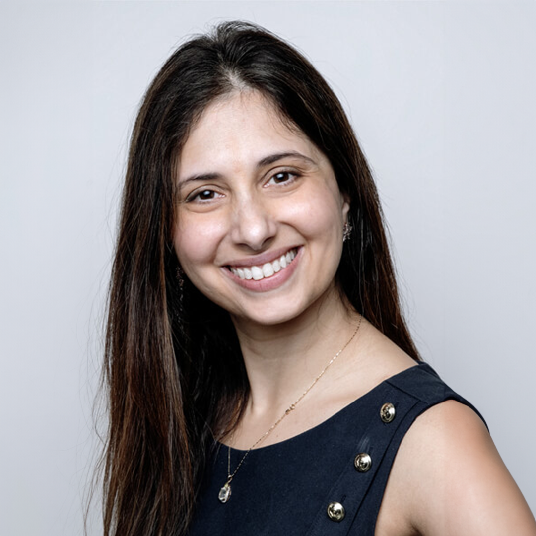 Close-up portrait of a woman with long dark brown hair, smiling, wearing a navy sleeveless top with silver button details and a delicate gold necklace, standing against a plain gray background.