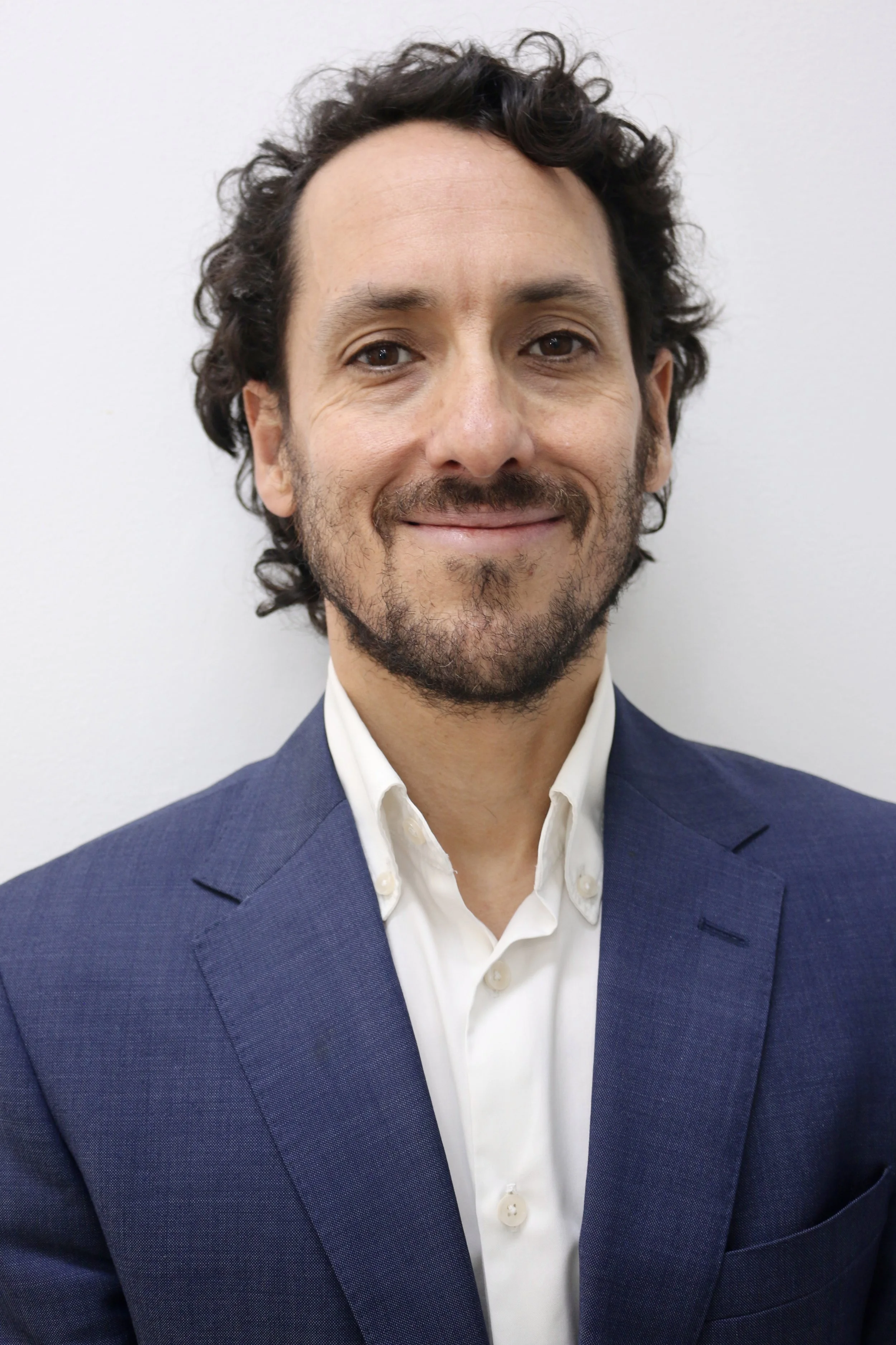 Portrait of a man with curly brown hair and a beard, wearing a navy blue suit and a white dress shirt, standing against a plain white background.