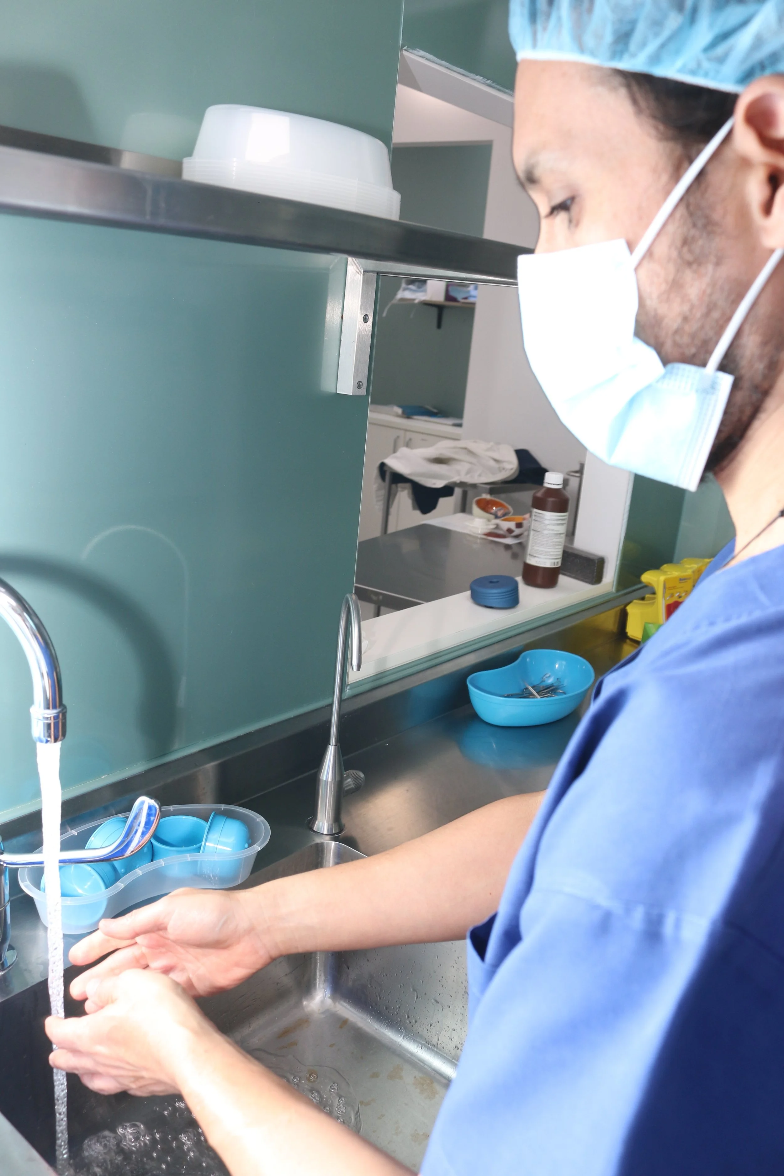 A healthcare worker wearing a face mask, hair cover, and scrubs is washing medical instruments in a sink filled with water in a hospital or clinic setting.