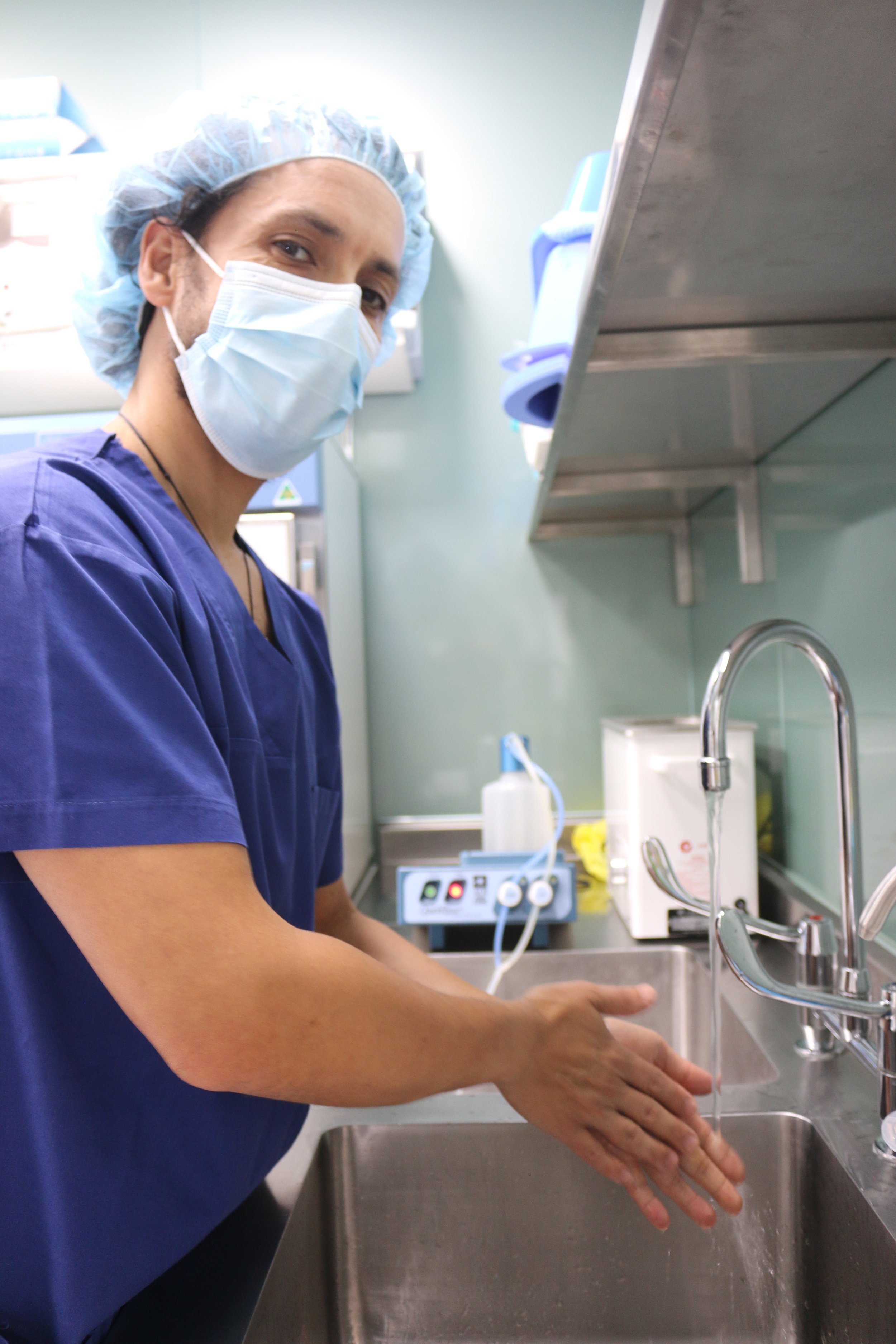 A healthcare professional washing their hands in a hospital or clinical setting. They are wearing a face mask, hair cover, and scrubs.