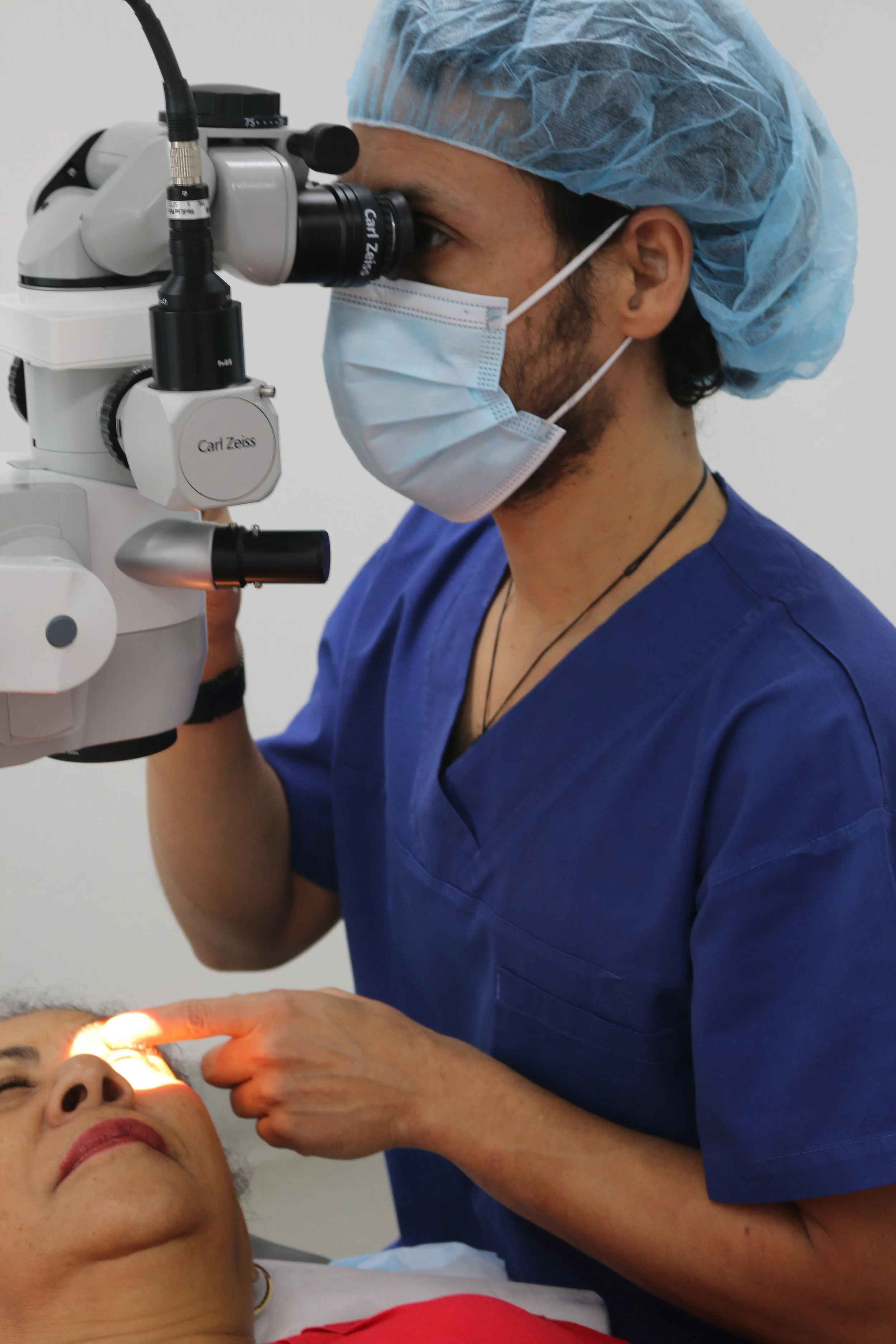 A medical professional in blue scrubs, a surgical mask, and a hairnet is examining a patient's eye using a slit lamp microscope in a clinical setting.