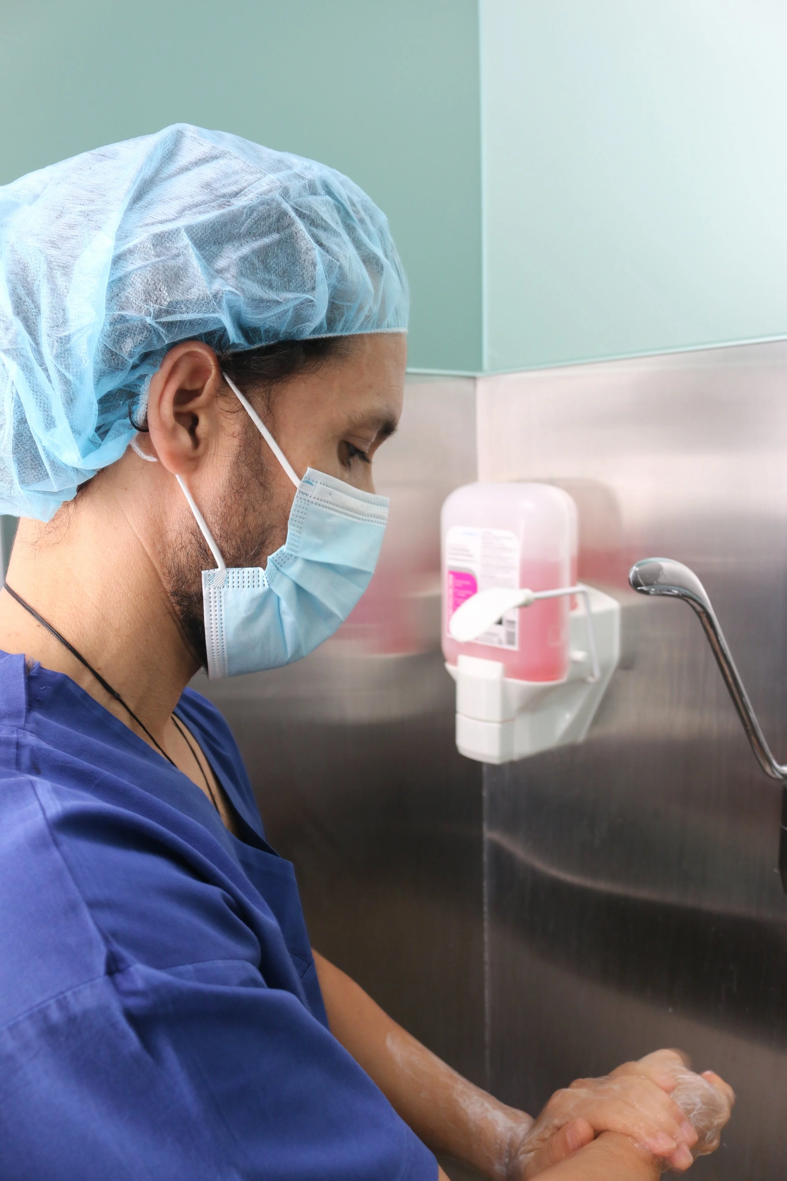 A healthcare worker wearing a blue scrub top, a disposable hair cap, and a face mask is washing their hands in a sink at a medical facility.