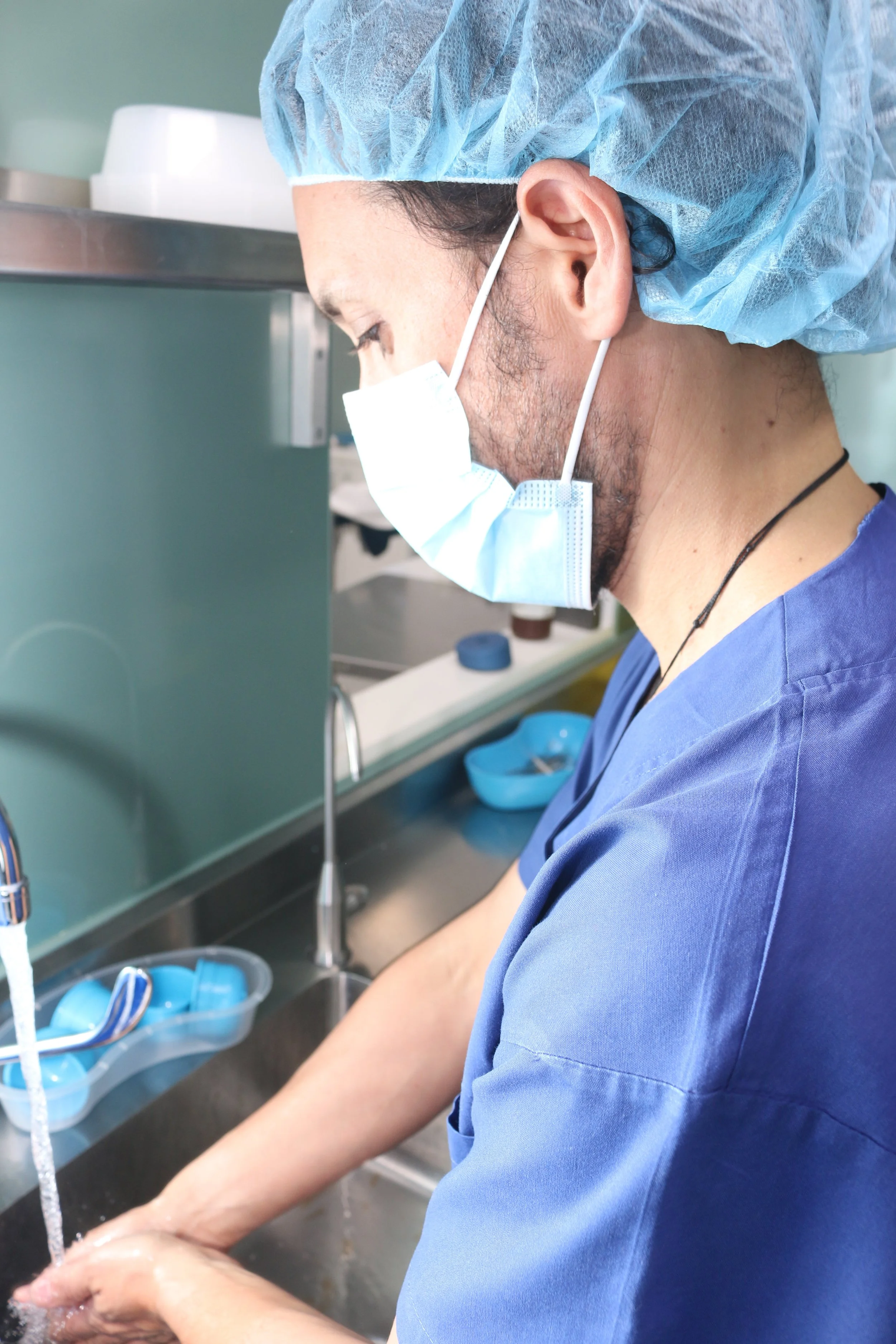 A healthcare professional wearing a blue scrubs, a disposable hair cap, and a face mask washing hands in a medical setting.