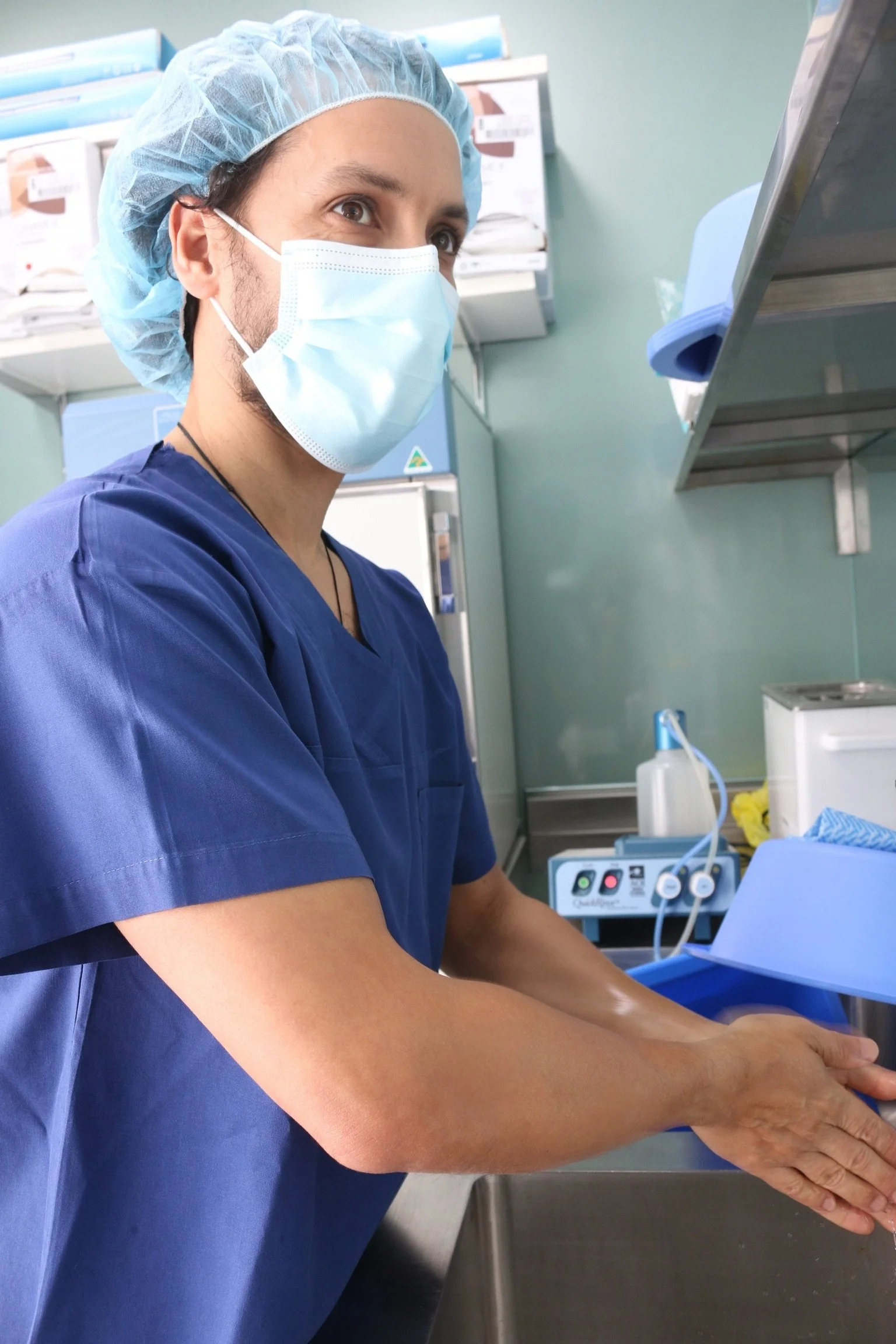 A healthcare professional wearing a blue scrub, surgical mask, and hair cap is preparing a patient for a medical procedure, with medical equipment in the background.