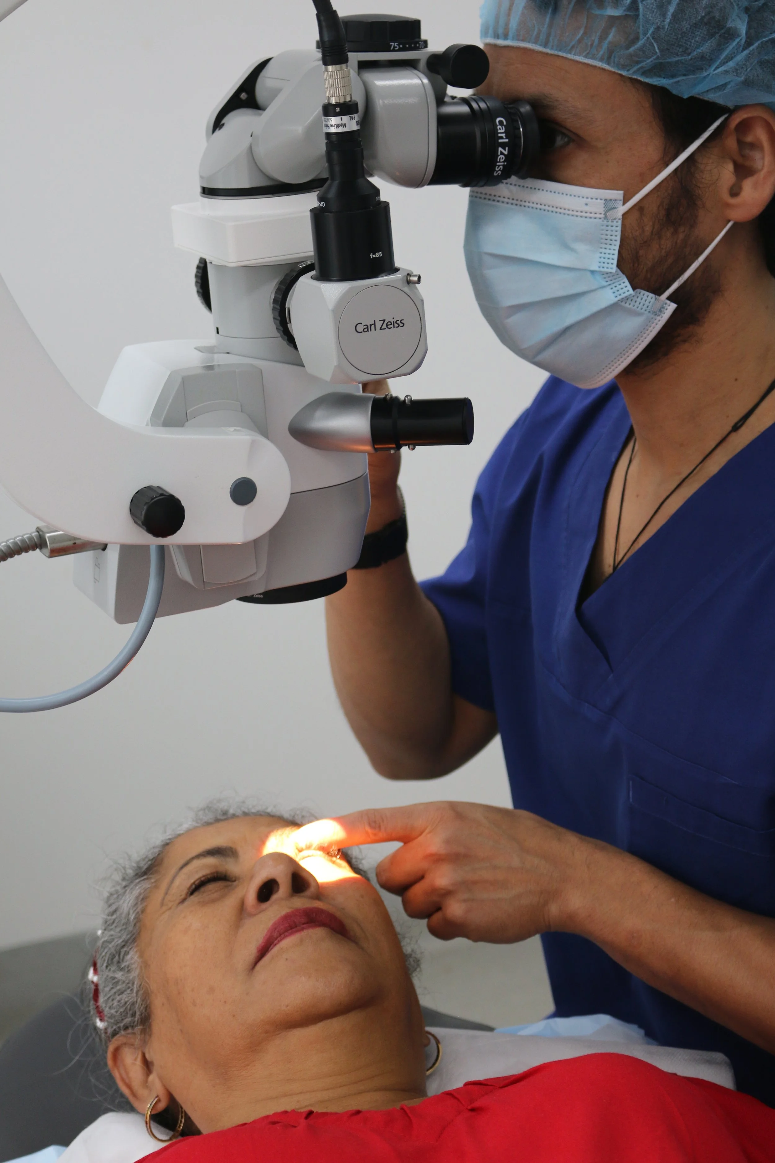 A healthcare professional using a surgical microscope to examine a patient lying down with closed eyes. The professional is wearing a blue scrub, a face mask, and a surgical cap. The patient's face, with a hand resting on her forehead, is illuminated