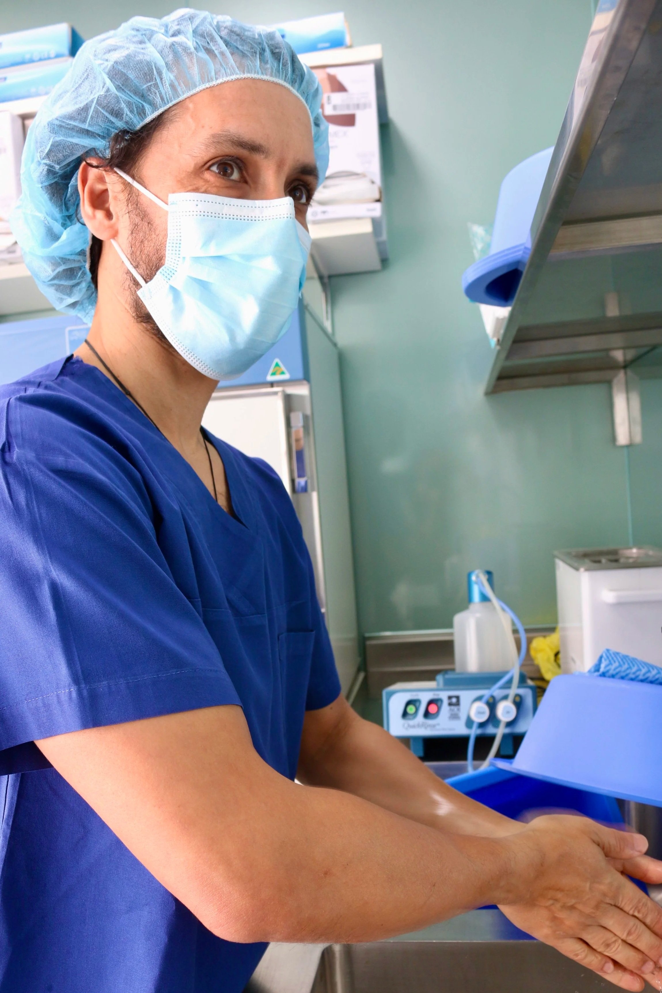 A healthcare professional in blue scrubs, wearing a surgical mask and hairnet, preparing to wash or sterilize their hands in a clinical setting with medical equipment and supplies in the background.