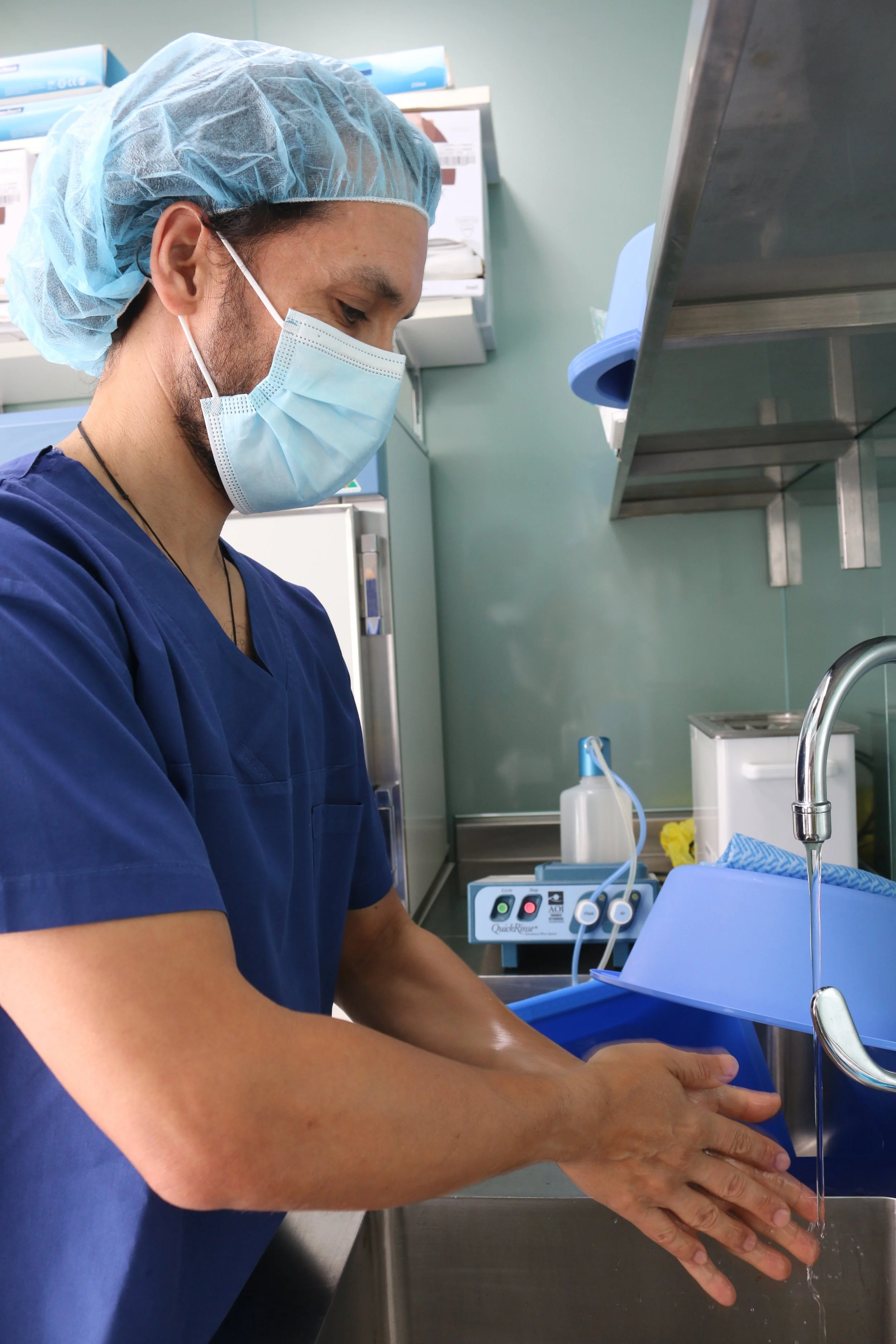 Medical professional in scrubs, surgical mask, and hair cap washing and sanitizing hands in a hospital or clinical setting.