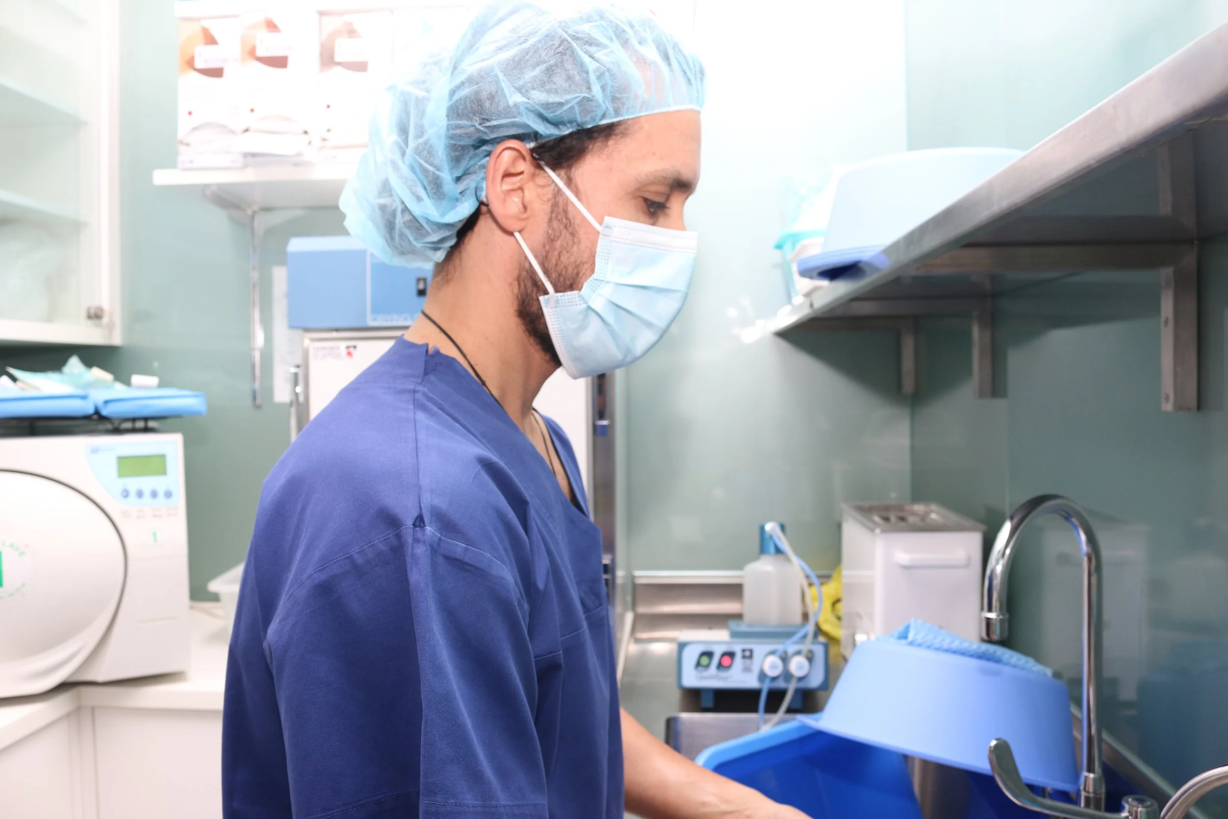 A healthcare professional, dressed in blue scrubs and a surgical mask, working in a medical or laboratory setting with scientific equipment and supplies around him.
