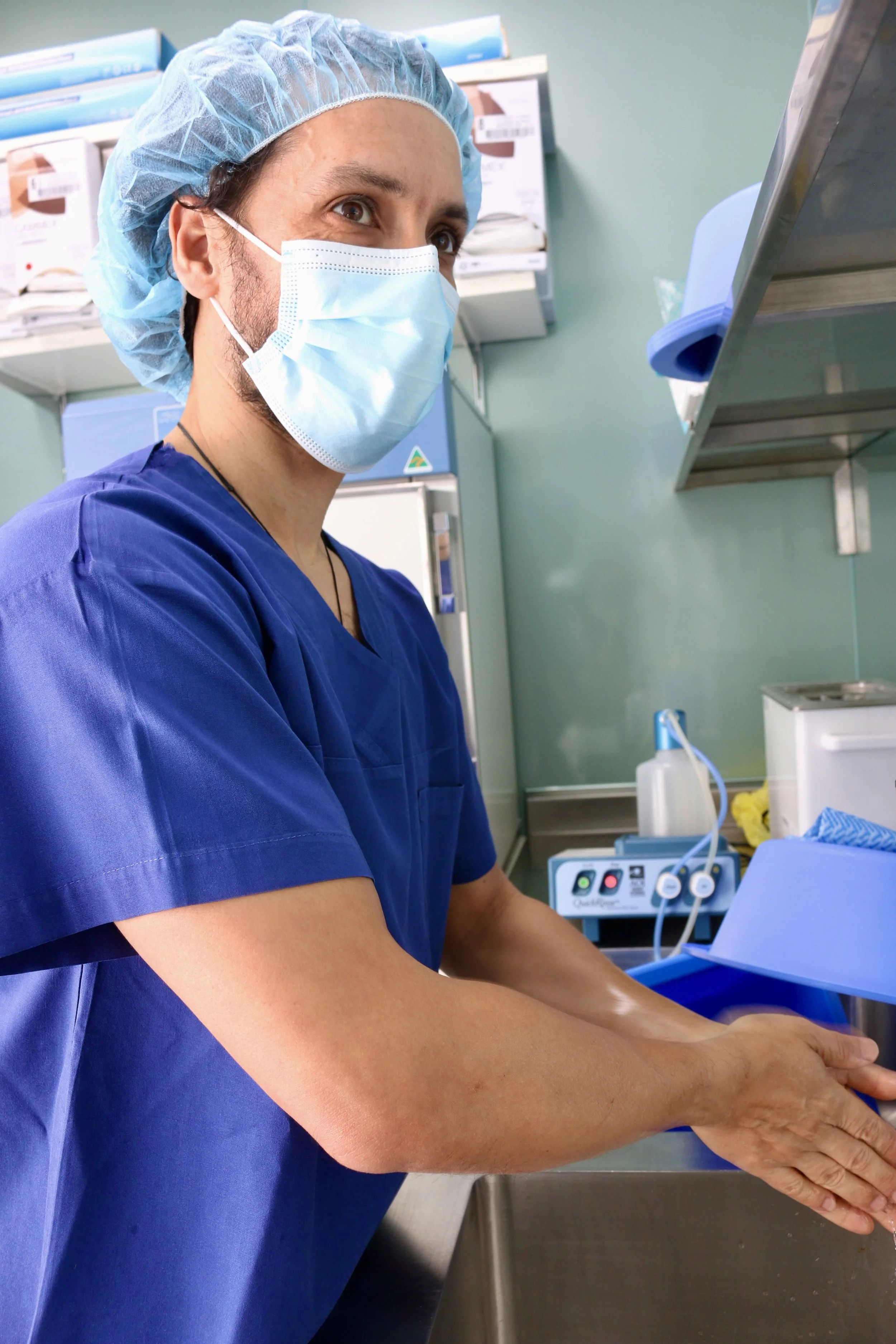 A healthcare worker wearing a blue surgical gown, a hairnet, and a face mask is preparing a patient for a medical procedure in a clinical setting.
