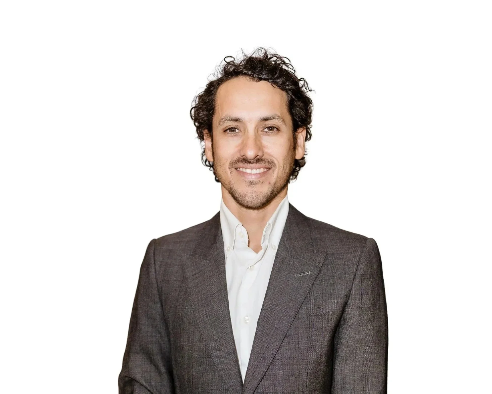 A man with curly dark hair and a beard, smiling slightly, dressed in a dark gray suit with a white shirt, standing against a plain white background.
