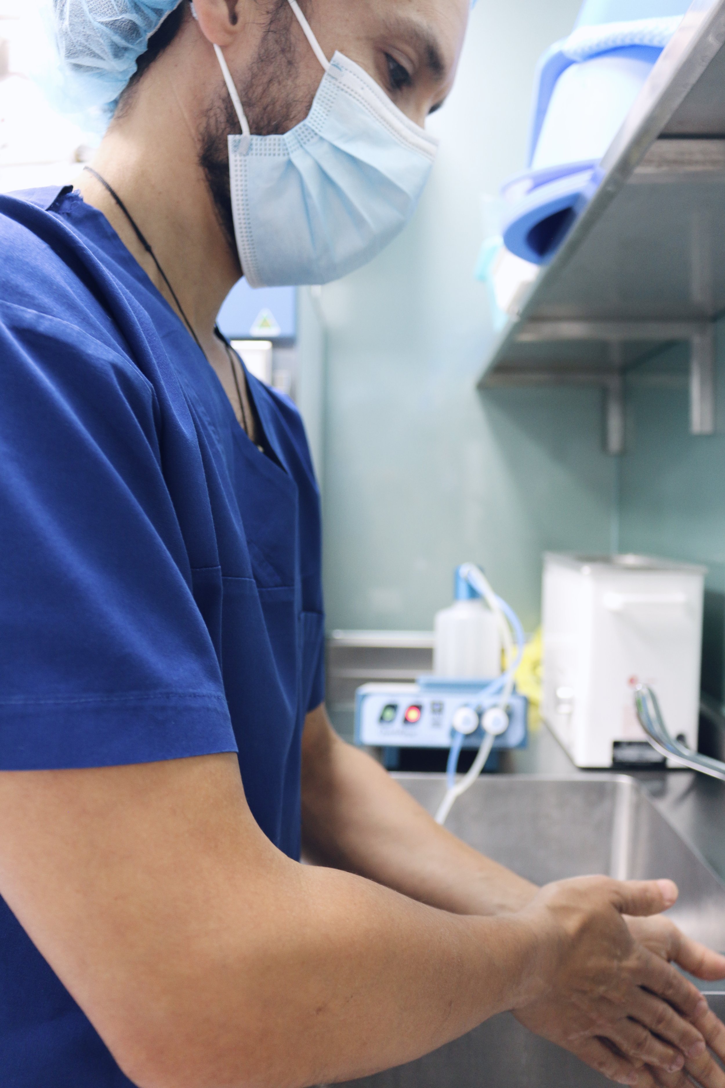 A healthcare worker wearing a blue uniform, surgical mask, and cap preparing for a procedure in a clinical setting, with medical equipment and supplies visible in the background.