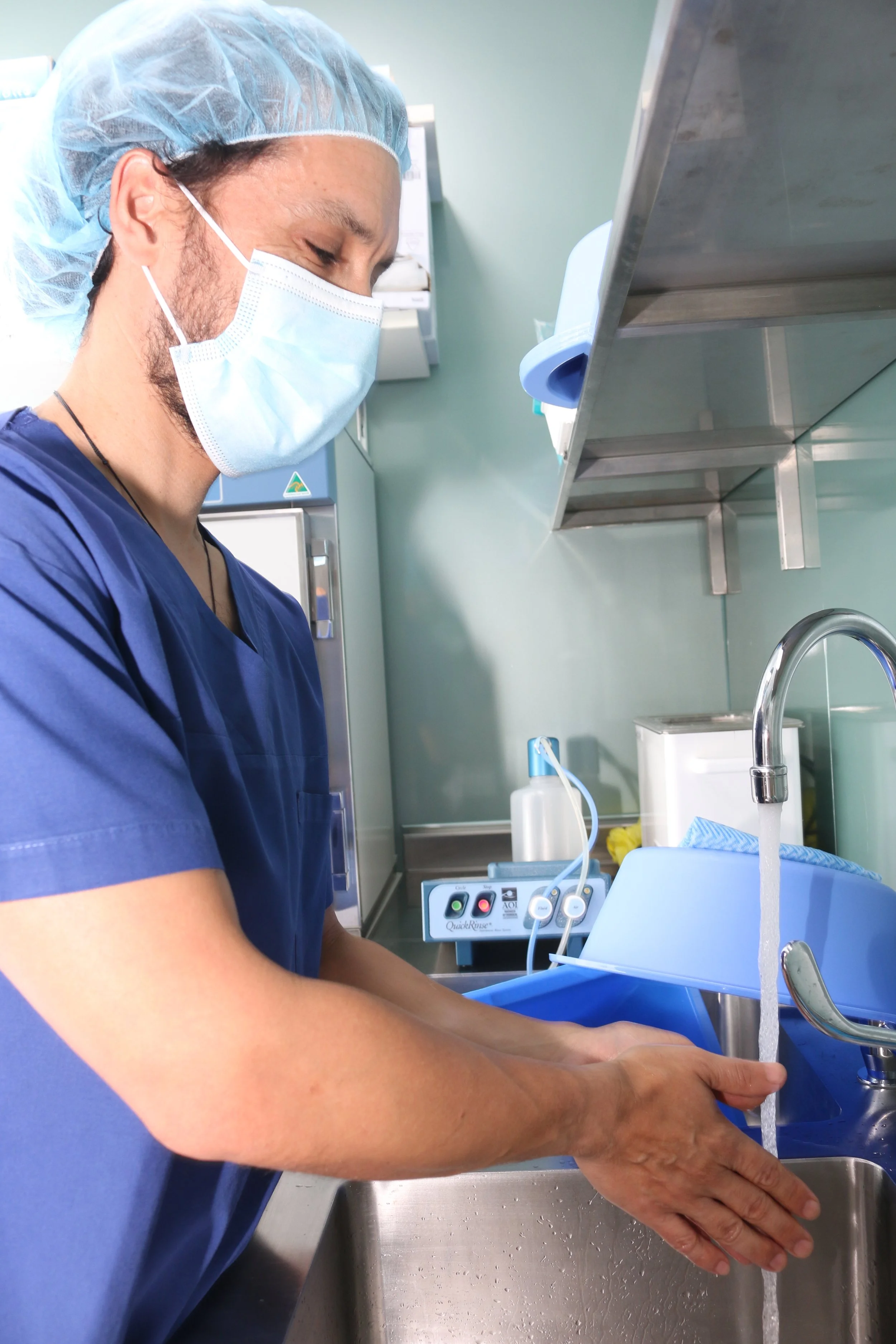 A healthcare professional in blue scrubs, face mask, and hair cover washing hands at a hospital sink.