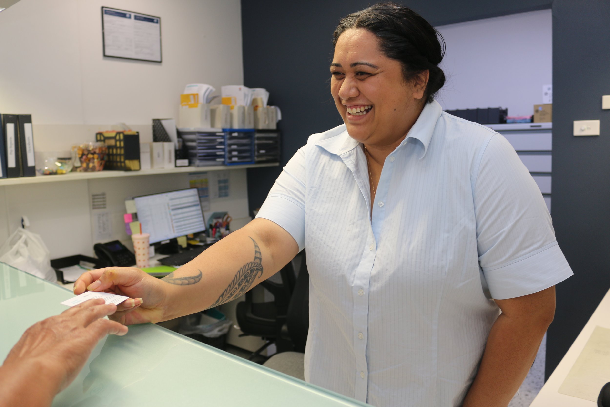 A woman with dark hair in a bun, wearing a white shirt, smiling and handing a card to another person at a reception desk in an office.