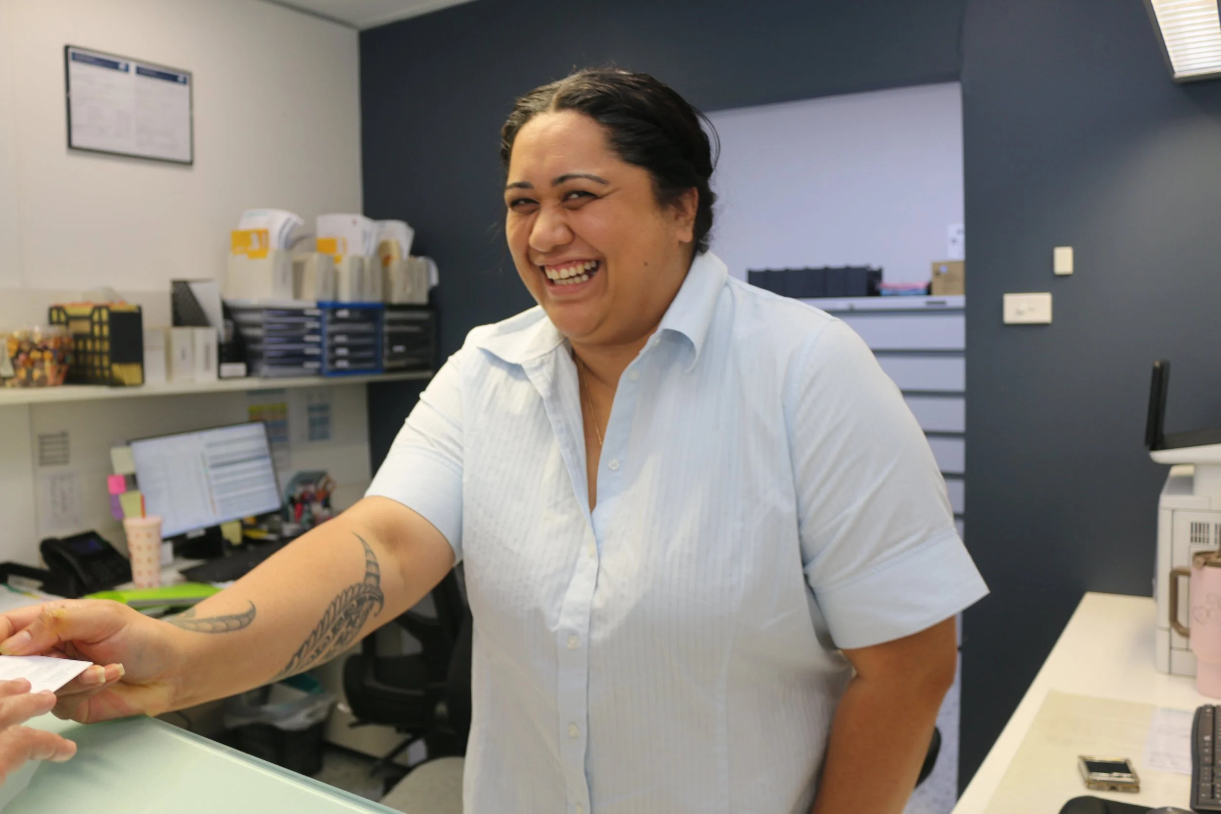 A woman with dark hair smiling and reaching out to receive a paper at an office desk.